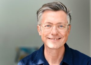 A middle-aged man with gray hair and clear eyeglasses smiles softly at the camera. He is wearing a navy blue shirt and is standing in front of a light, blurred background. at Hochschule Coburg