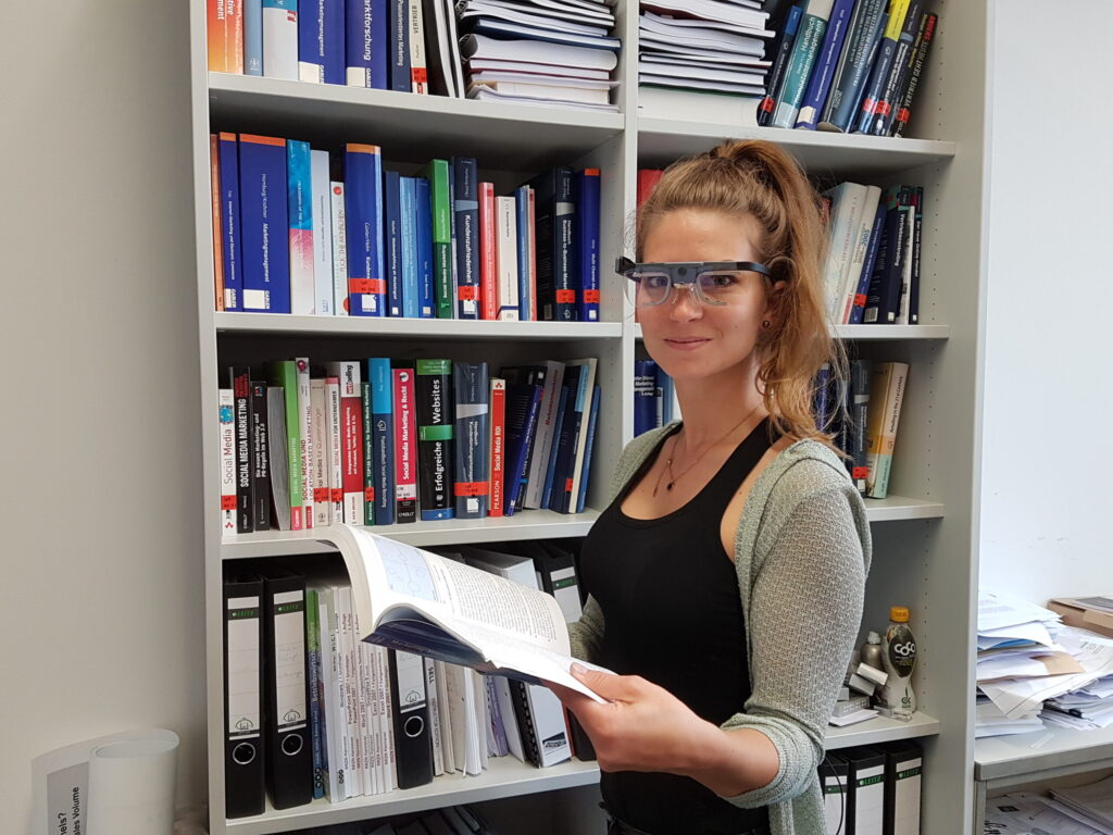 A woman wearing smart glasses stands in front of a bookshelf filled with books at Hochschule Coburg, holding an open book and smiling at the camera. She is dressed casually and appears to be in an office or library setting. at Hochschule Coburg