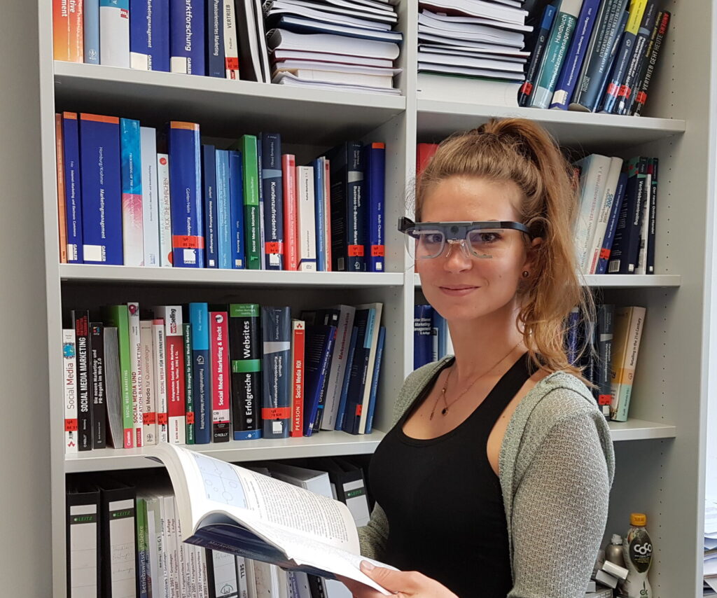 Eine Frau mit einer Eye-Tracking-Brille steht vor einem mit Büchern und Ordnern gefüllten Bücherregal an der Hochschule Coburg, hält ein aufgeschlagenes Buch in der Hand und lächelt in die Kamera in einer Büroumgebung. at Hochschule Coburg