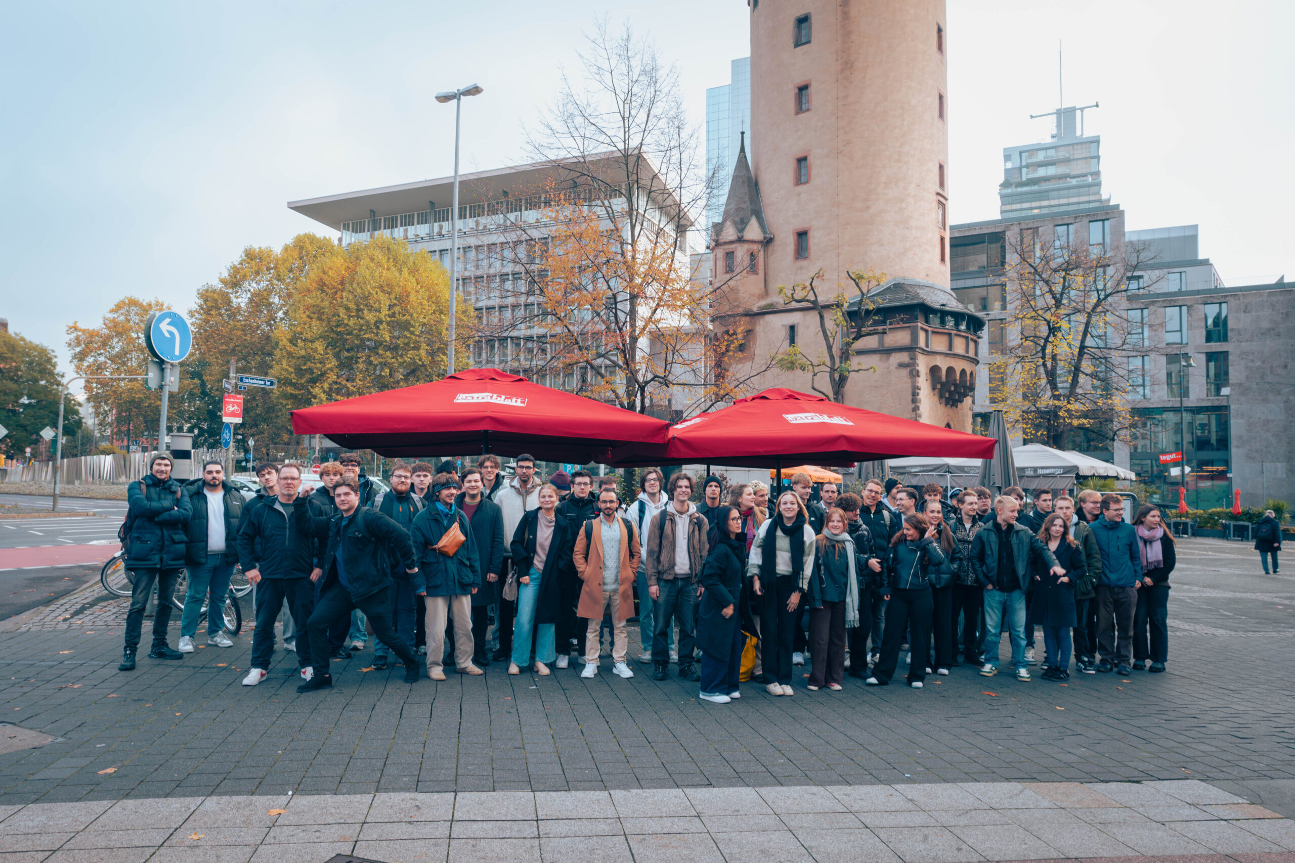 Eine große Gruppe von Menschen posiert gemeinsam im Freien auf einer Straße in der Stadt, mit zwei roten Regenschirmen im Hintergrund und modernen und historischen Gebäuden an einem bewölkten Tag - vielleicht Studenten oder Fachleute aus der Welt der Wirtschaftsinformatik. at Hochschule Coburg