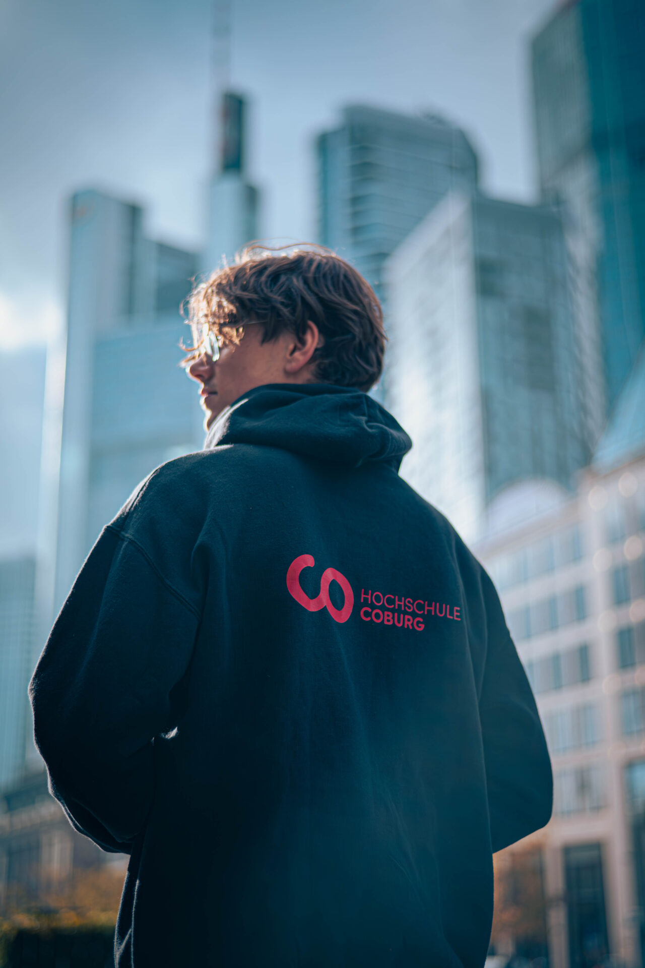 Eine Person, die einen schwarzen "Hochschule Coburg"-Kapuzenpulli mit rotem Logo trägt, steht im Freien in einer Stadt mit hohen modernen Gebäuden - vielleicht ein Symbol für neue Horizonte in der Wirtschaftsinformatik oder der Finanzwelt. Die Person blickt weg, schaut zur Seite. at Hochschule Coburg