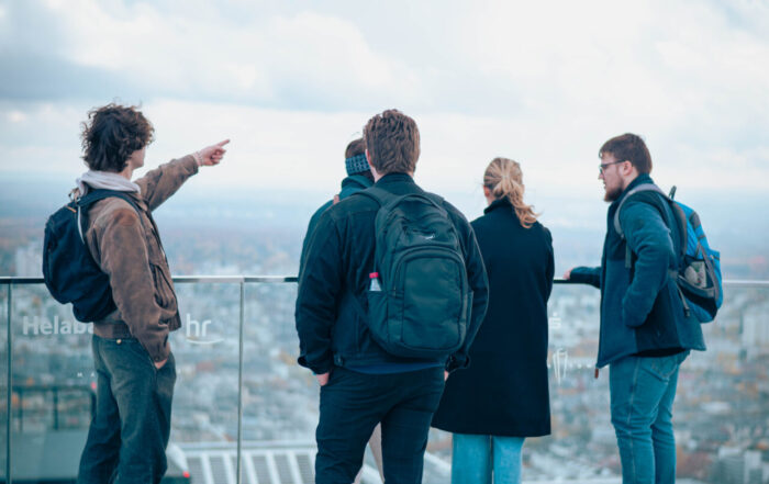 Fünf Personen stehen auf einer Dachterrasse mit Blick über die Stadt. Einer zeigt in die Ferne, als würde er der Gruppe eine Theorie aus der Wirtschaftsinformatik erklären. Der Himmel ist bewölkt, und alle tragen Jacken oder Mäntel. at Hochschule Coburg