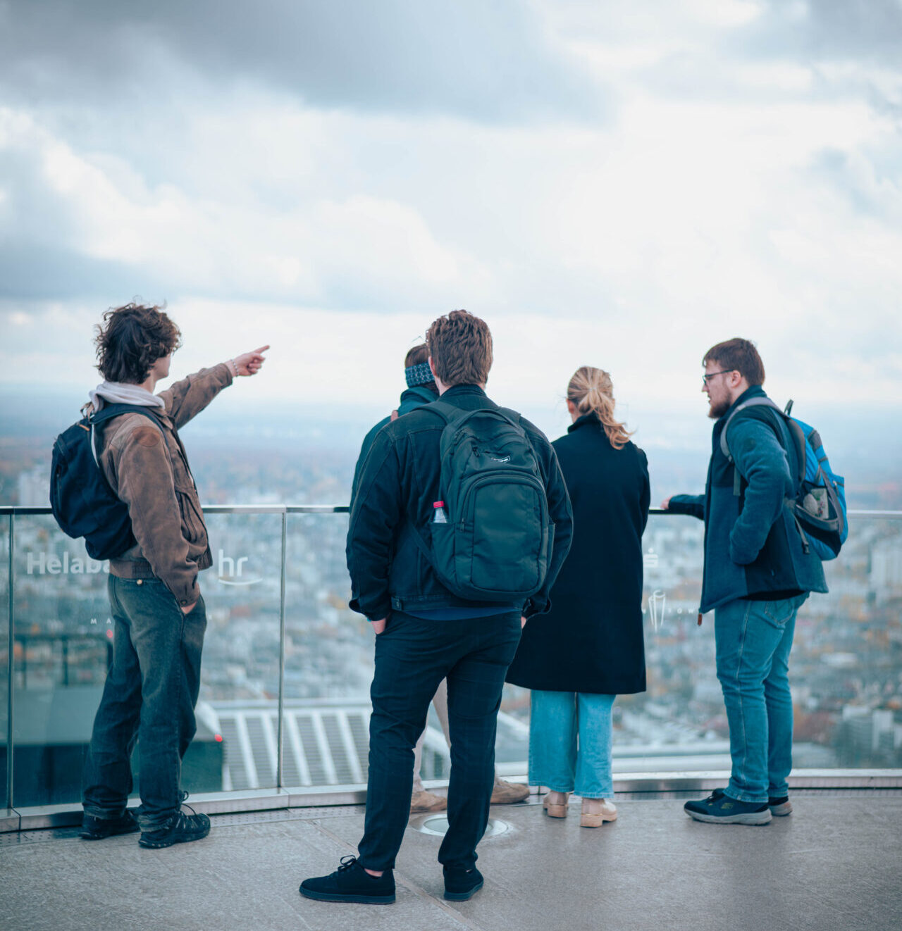 Fünf Personen stehen auf einer Dachterrasse mit Blick über die Stadt. Einer zeigt in die Ferne, als würde er der Gruppe eine Theorie aus der Wirtschaftsinformatik erklären. Der Himmel ist bewölkt, und alle tragen Jacken oder Mäntel. at Hochschule Coburg
