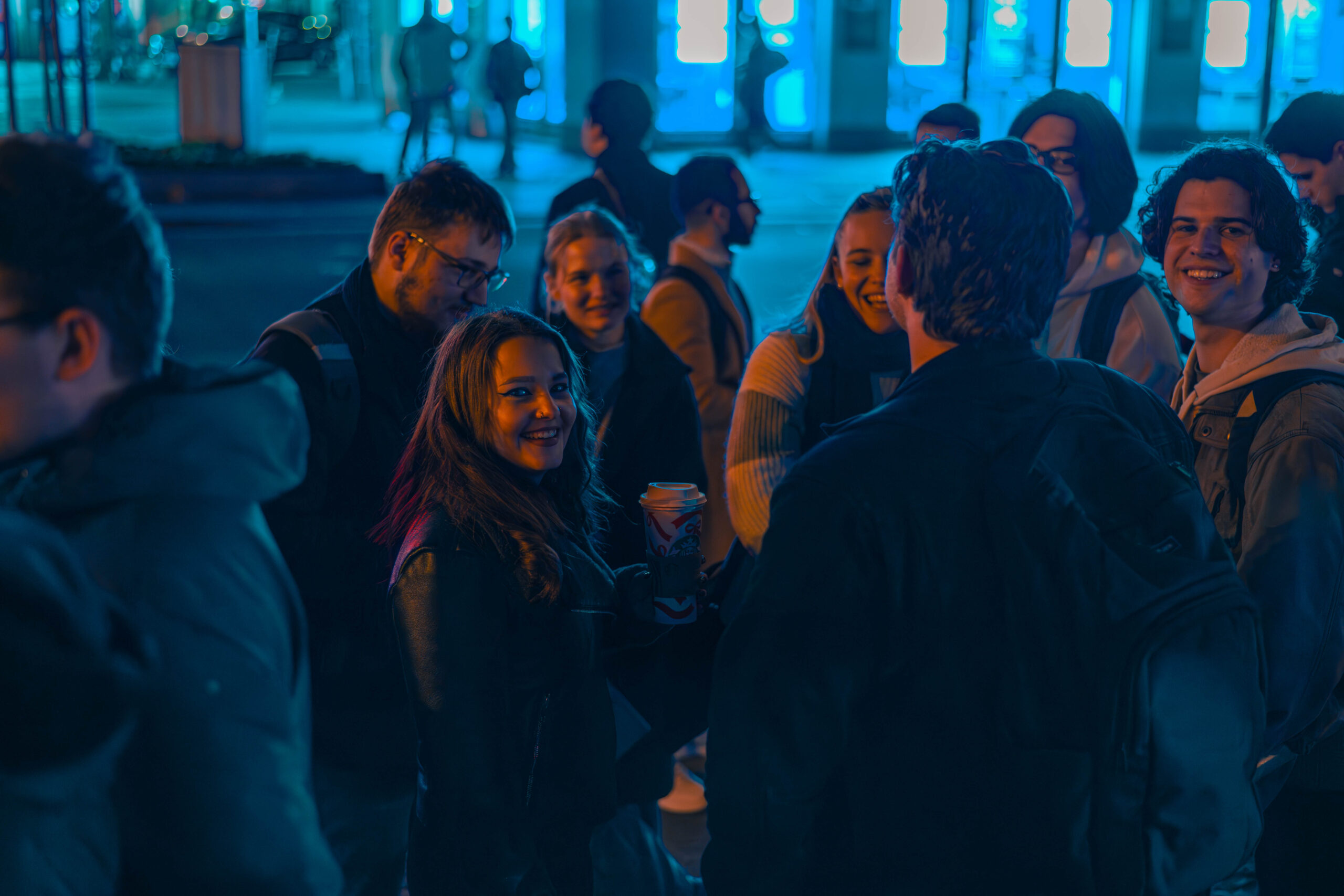 Eine Gruppe junger Erwachsener versammelt sich nachts draußen, lächelt und unterhält sich unter blauen Neon-Straßenlaternen. Eine Person in der Mitte hält ein Getränk in der Hand und gibt Einblicke in die Wirtschaftsinformatik, während alle die lebhafte Unterhaltung genießen. at Hochschule Coburg