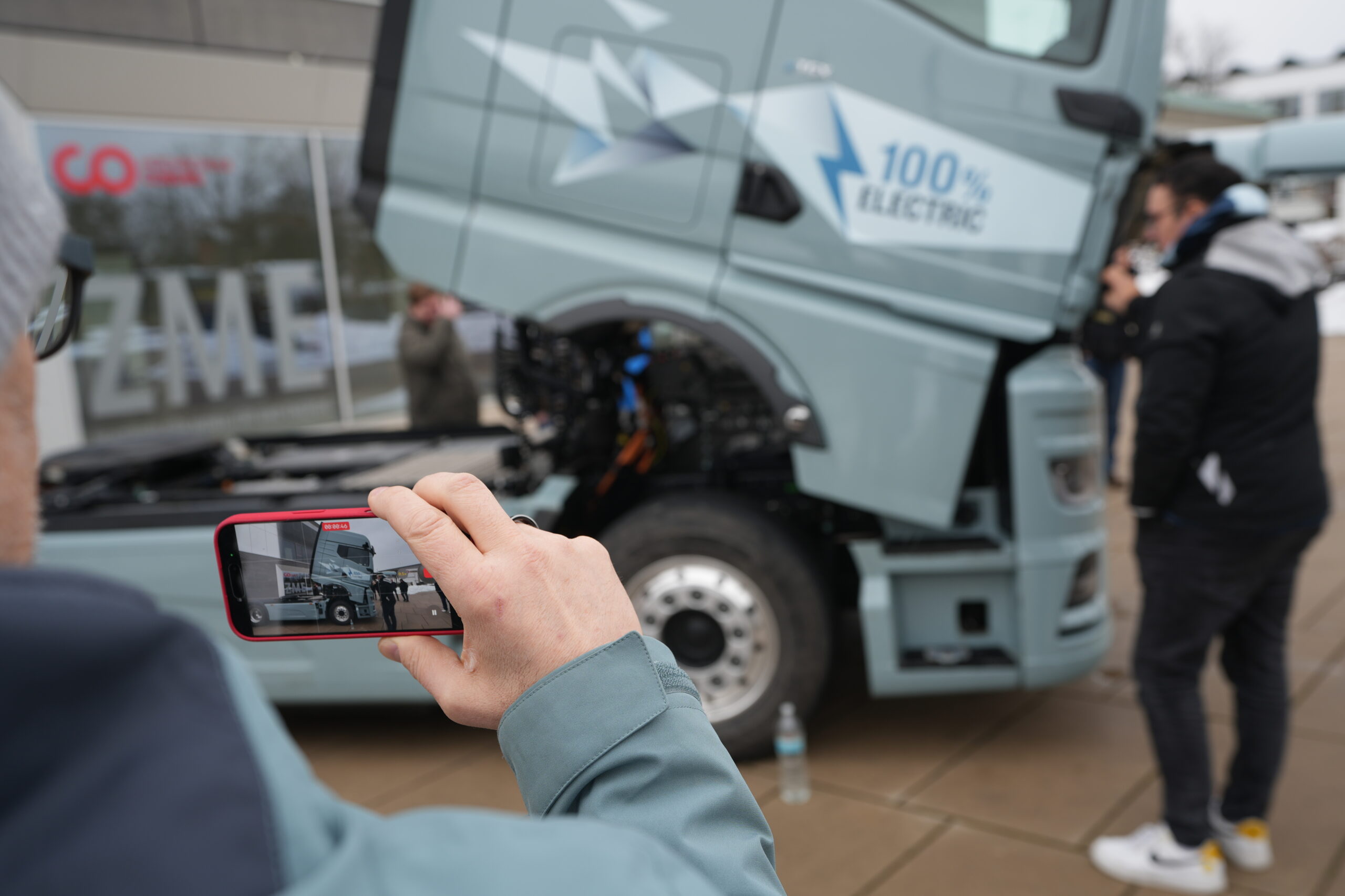 Eine Person fotografiert mit ihrem Smartphone einen großen Elektro-LKW mit geöffneter Motorhaube, auf der "100% ELECTRIC" steht. In der Nähe steht jemand bereit, um über E-Mobilität und Weiterbildungsmöglichkeiten für Berufsschulen zu sprechen. at Hochschule Coburg