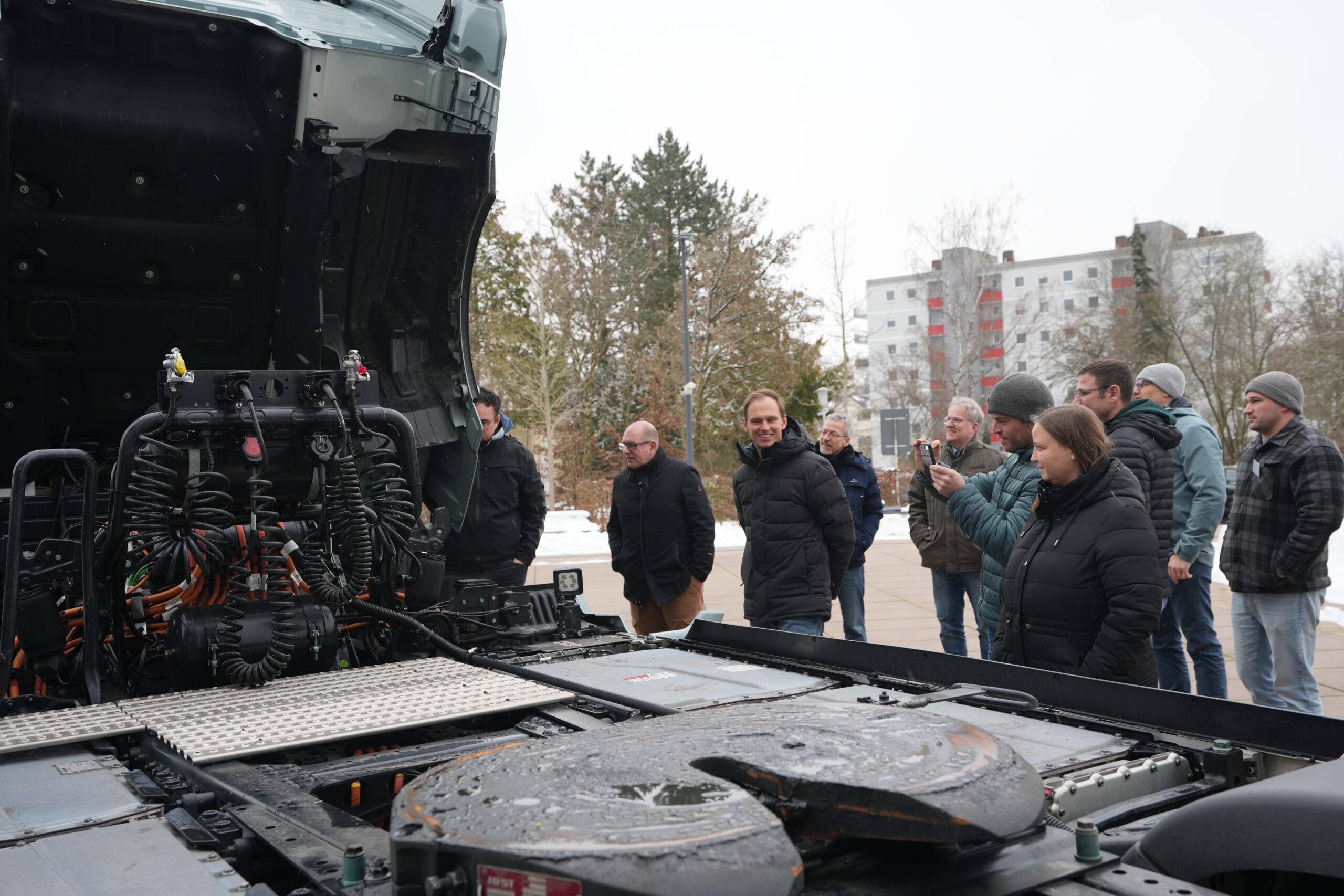 Eine Gruppe von Personen in Winterkleidung steht im Freien und beobachtet und fotografiert den freiliegenden Motor und die Maschinen eines großen Lastkraftwagens mit angehobenem Fahrerhaus - möglicherweise Teil einer Weiterbildung zum Thema E-Mobilität für Berufsschulen. Im Hintergrund sind Schnee und Gebäude zu sehen. at Hochschule Coburg