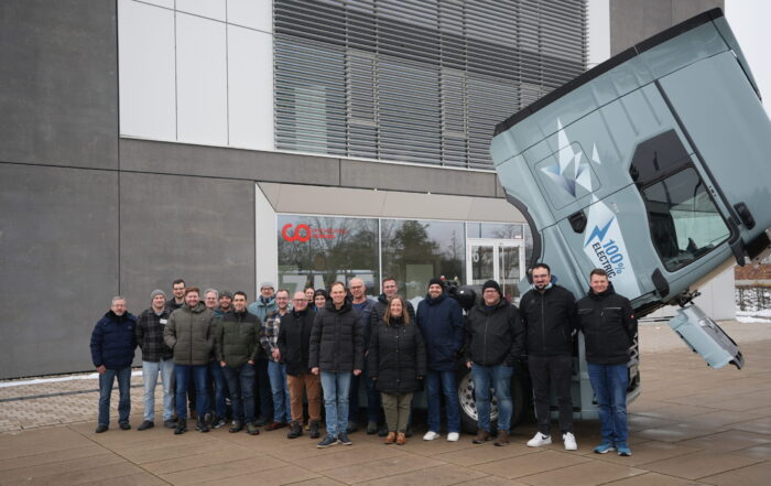 Eine Gruppe von Menschen in Winterkleidung steht vor einem modernen Gebäude und einem großen Lastwagen mit nach vorne gekipptem Fahrerhaus, möglicherweise im Rahmen einer von den örtlichen Berufsschulen organisierten E-Mobilitäts-Weiterbildung. Der Boden ist nass und am Rand liegt etwas Schnee. at Hochschule Coburg