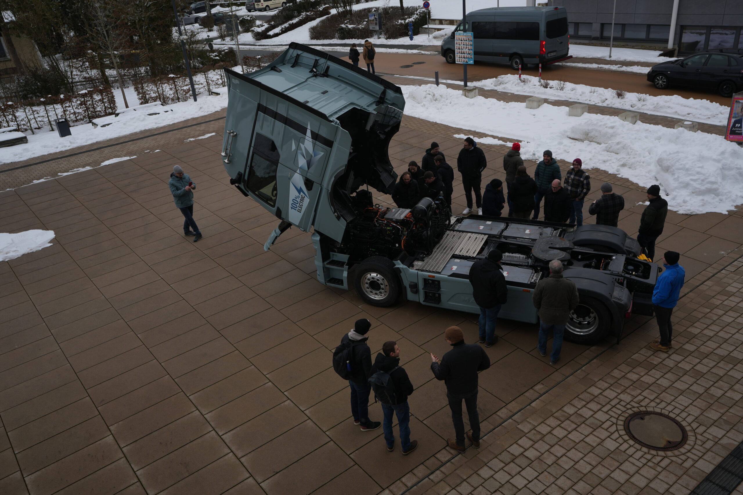 Eine Gruppe von Menschen steht um einen großen Lastwagen mit offener Motorhaube, der auf einer verschneiten, gepflasterten Fläche geparkt ist - einige machen Fotos. Die Szene deutet auf eine Weiterbildungsveranstaltung zum Thema E-Mobilität für Berufsschulen hin, denn am Rande des Gehwegs türmt sich der Schnee. at Hochschule Coburg