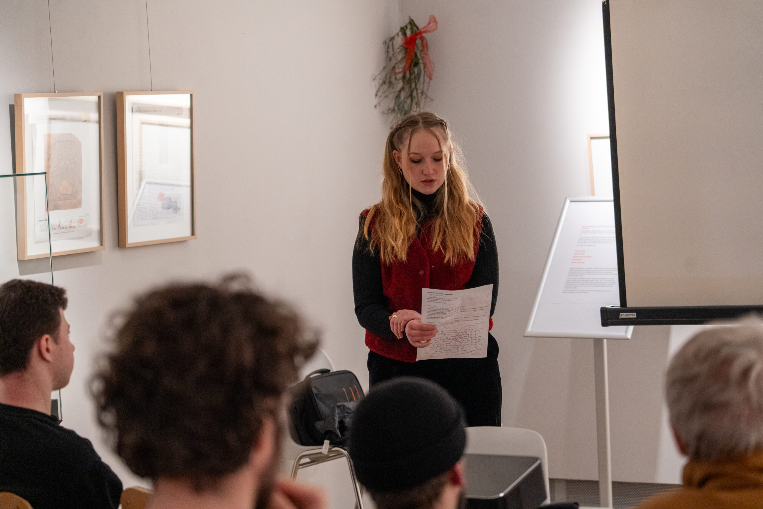 Eine Frau steht am Eingang eines kleinen Raums, hält Papiere in der Hand und spricht zu einem Publikum über mögliche Zukünfte. An den Wänden hängen gerahmte Kunstwerke, und neben ihr ist teilweise eine Projektionsfläche zu sehen. at Hochschule Coburg