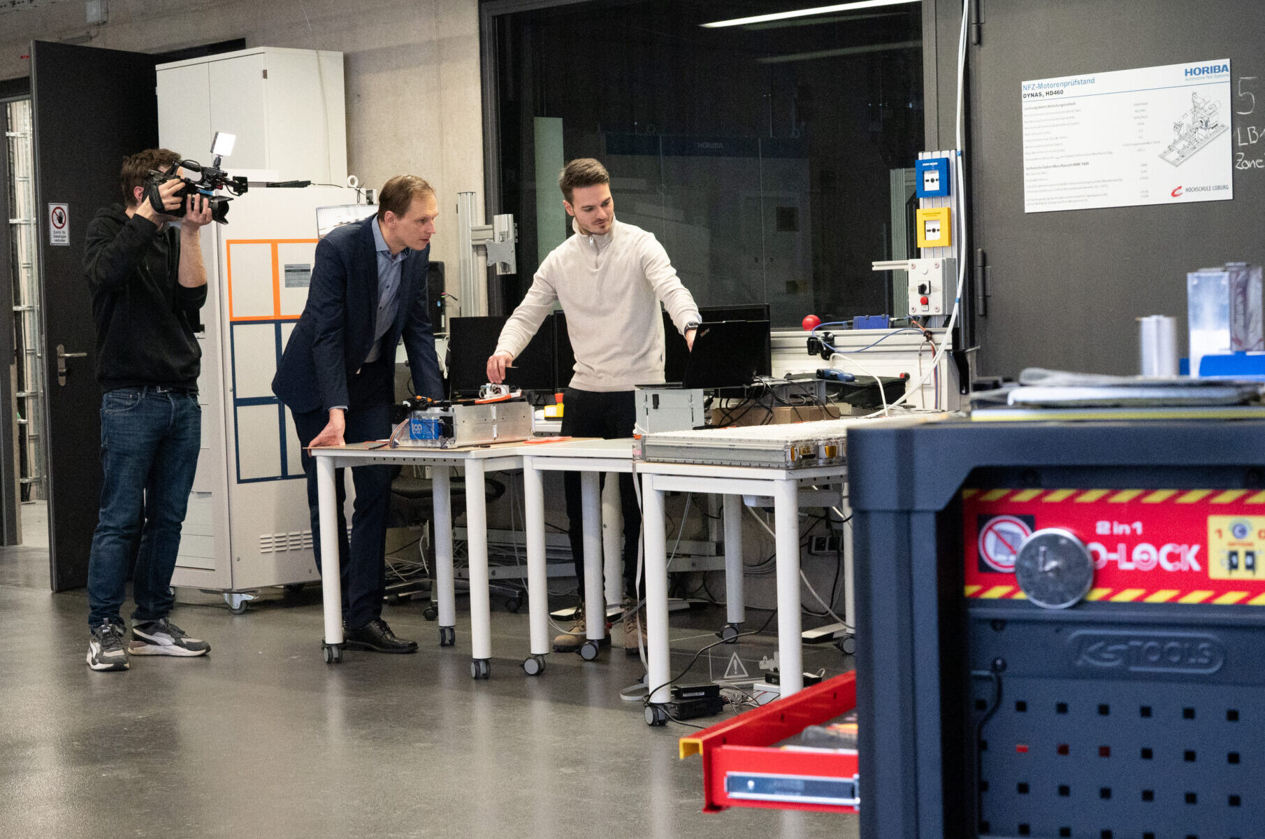 Zwei Männer arbeiten an einem Tisch mit elektronischen Geräten in einem modernen Industrielabor für Coburger Batterieforschung und E-Mobilität, während ein Kameramann sie filmt. Der Raum ist mit Werkzeugen, Schränken und einem geparkten weißen Auto ausgestattet, das teilweise sichtbar ist. at Hochschule Coburg