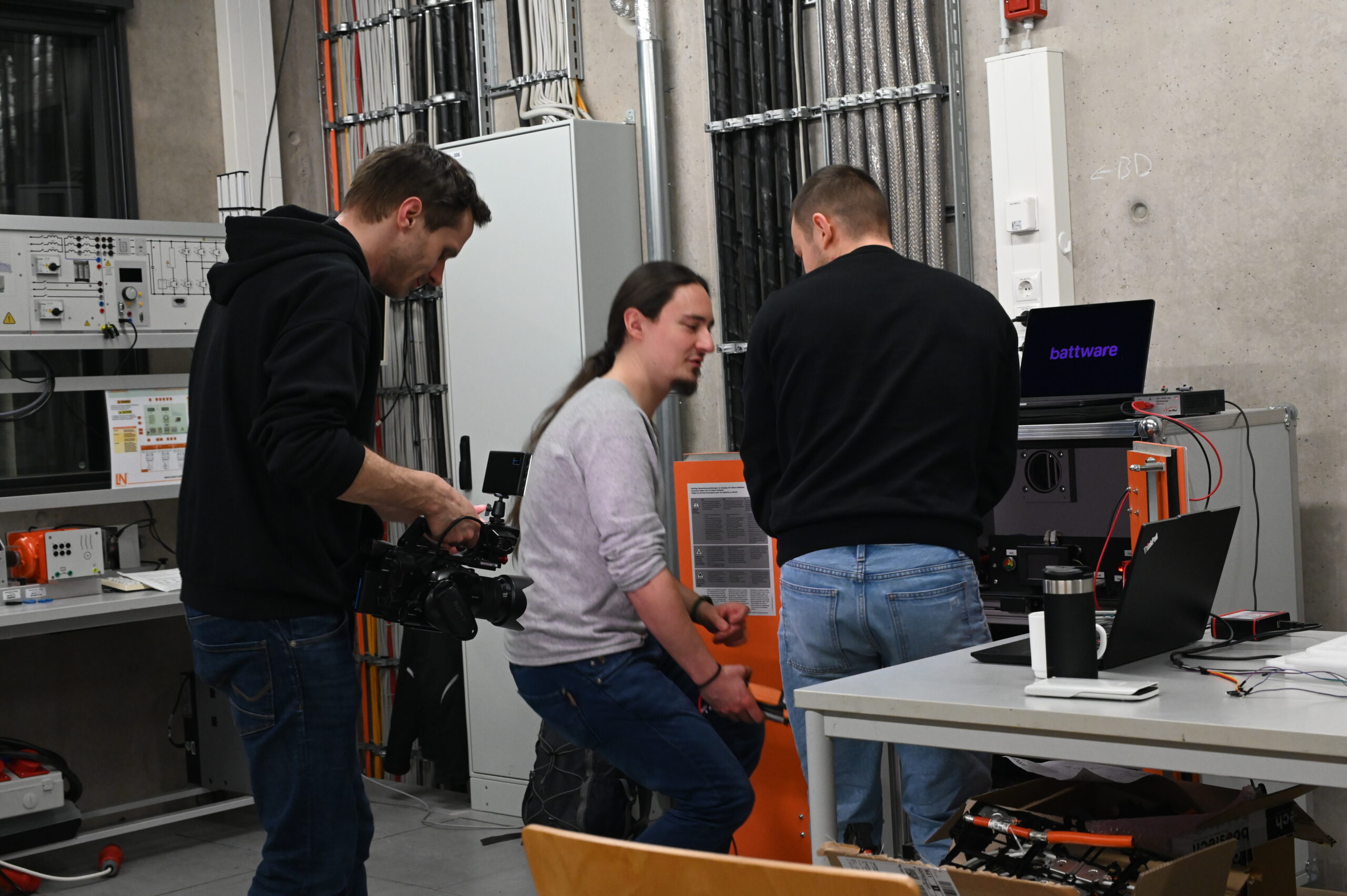 Drei Personen arbeiten in einem Labor mit technischen Geräten; eine hält eine Kamera, eine andere justiert ein Gerät, die dritte steht an einem Tisch mit Laptops und Kabeln. Auf einem Monitor ist "batteware" zu sehen, ein Hinweis auf die Coburger Batterieforschung in E-Mobilität. at Hochschule Coburg