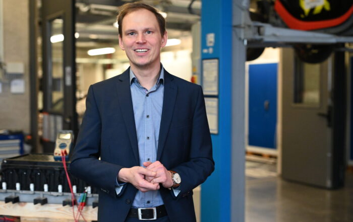 Ein Mann im Anzug steht lächelnd in einer modernen Industrie- oder Laborumgebung, die sich auf die Coburger Batterieforschung konzentriert, mit blauen Wänden, Geräten und fortschrittlichen Maschinen im Hintergrund. at Hochschule Coburg