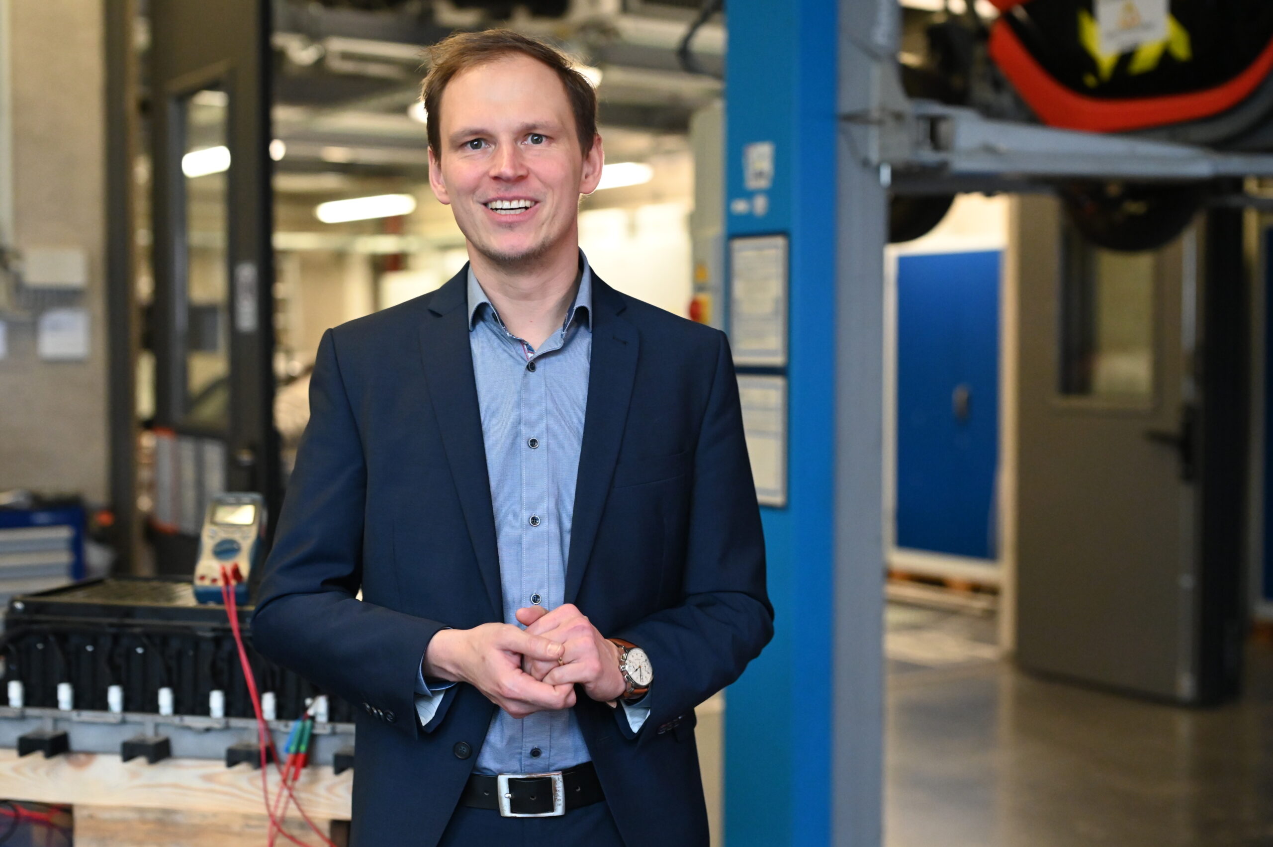 Ein Mann im Anzug steht lächelnd in einer modernen Industrie- oder Laborumgebung, die sich auf die Coburger Batterieforschung konzentriert, mit blauen Wänden, Geräten und fortschrittlichen Maschinen im Hintergrund. at Hochschule Coburg