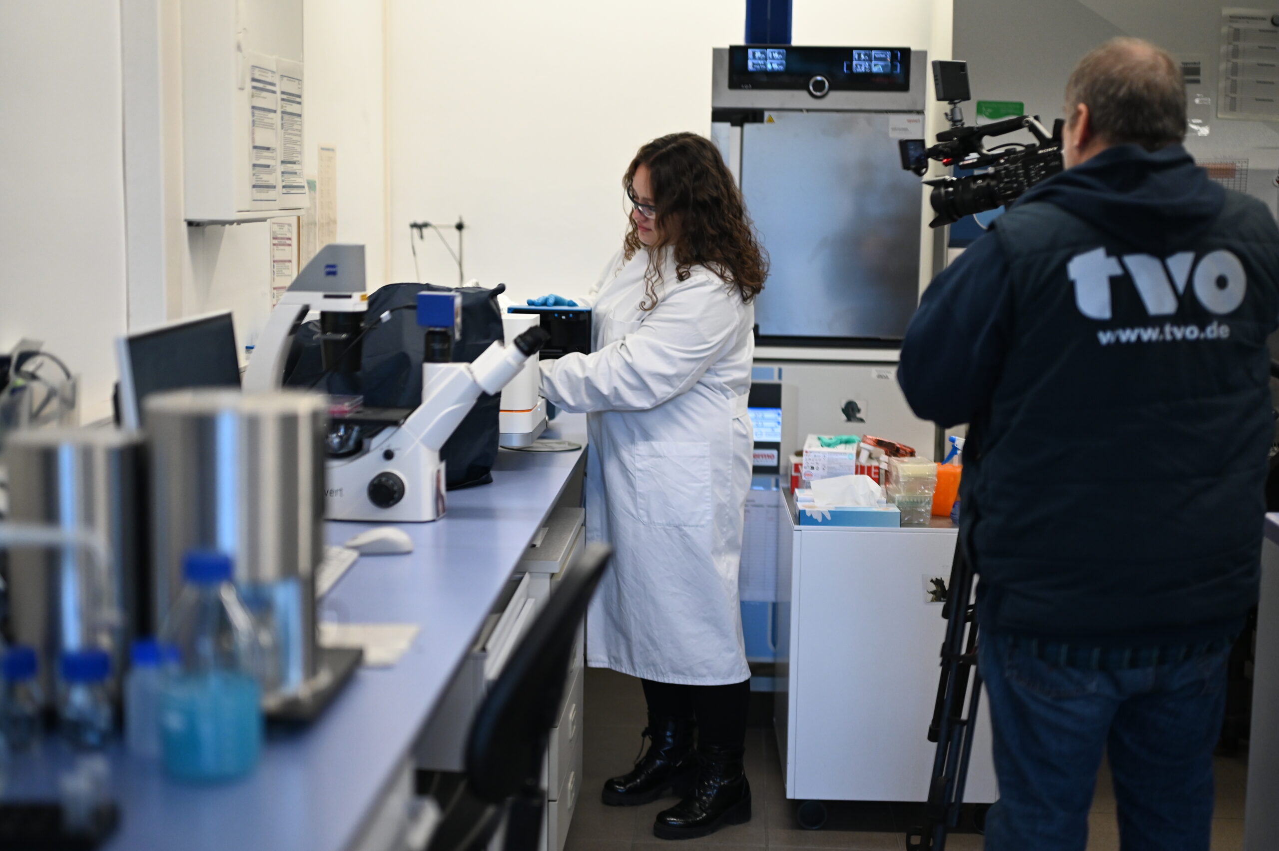 Eine Frau in einem weißen Laborkittel arbeitet mit wissenschaftlichen Geräten in einem Labor, das aktuelle Forschung zur Biologie der Verjüngung erforscht, während ein Kameramann in einer "tvo.de"-Jacke sie filmt. Auf den Theken sind verschiedene Laborutensilien und Geräte zu sehen. at Hochschule Coburg