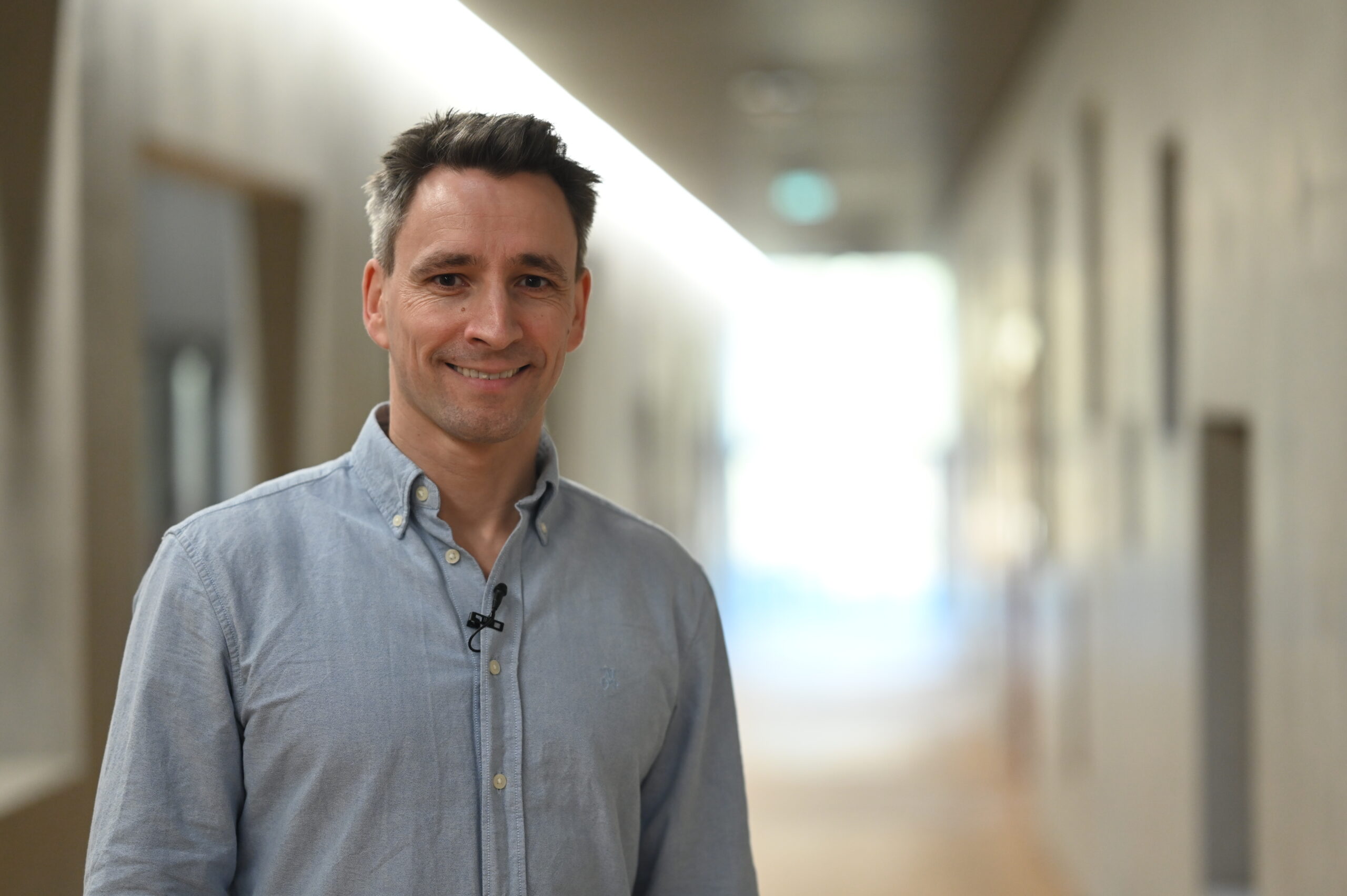 Ein Mann in einem hellblauen Button-up-Hemd steht in einem gut beleuchteten Hausflur und lächelt in die Kamera. Er hat kurzes dunkles Haar, ein kleines Mikrofon an seinem Hemd befestigt und scheint bereit für einen TV-Beitrag über aktuelle Forschung zur Biologie der Verjüngung. at Hochschule Coburg