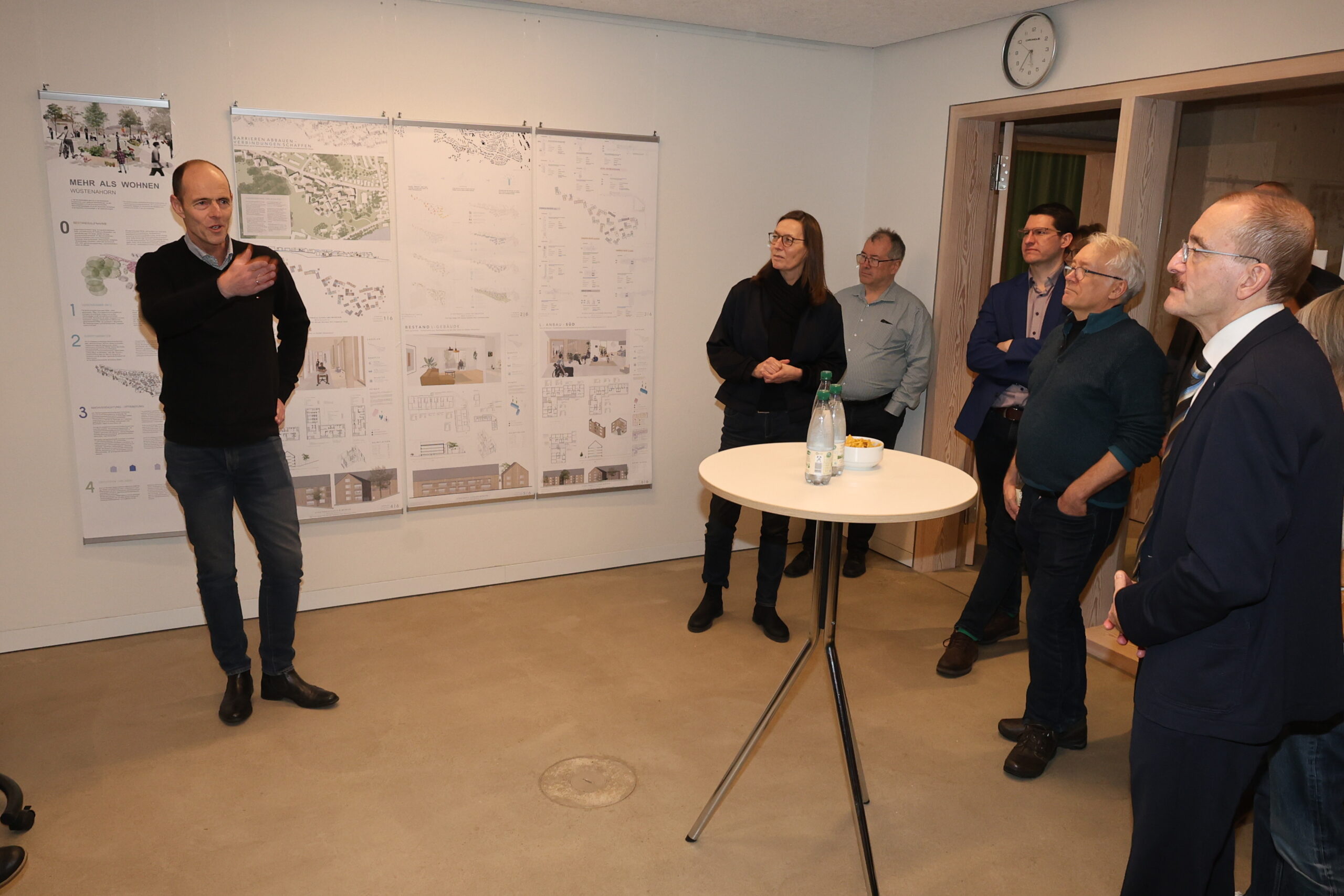 Eine Gruppe von Menschen steht in einem Raum und hört einem Mann zu, der Architekturpläne für eine Nachkriegssiedlung präsentiert, die an der Wand hängen. In der Mitte steht ein runder Tisch mit Flaschen und Gläsern, der die Atmosphäre der Ausstellung noch verstärkt. at Hochschule Coburg