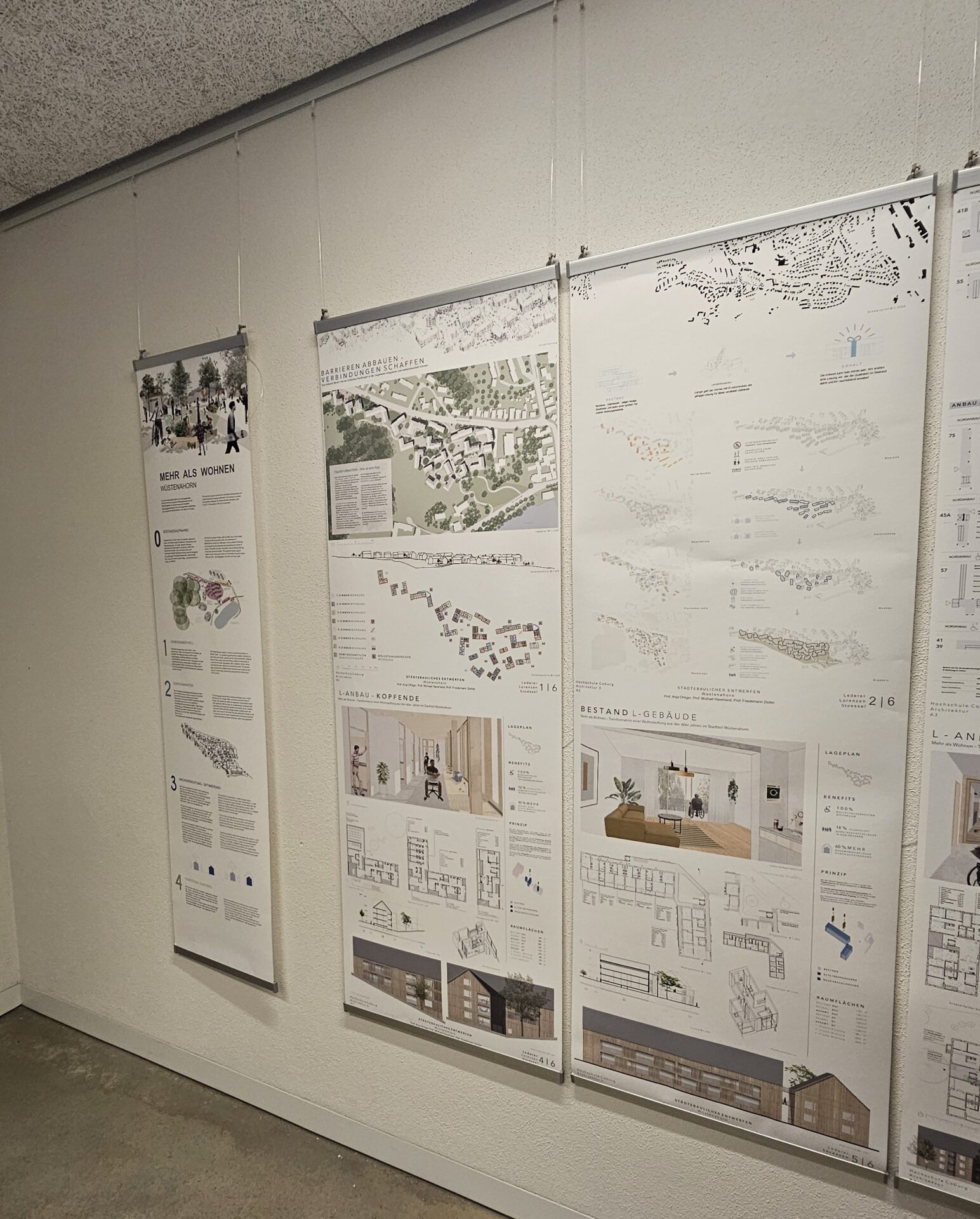 Vier große vertikale Plakate mit architektonischen Plänen, Diagrammen und Texten werden in einer Ausstellung mit Drähten und Haken an einer weißen Wand aufgehängt und bieten neue Perspektiven für die Gestaltung von Nachkriegssiedlungen in einem Flur oder einer Galerie. at Hochschule Coburg