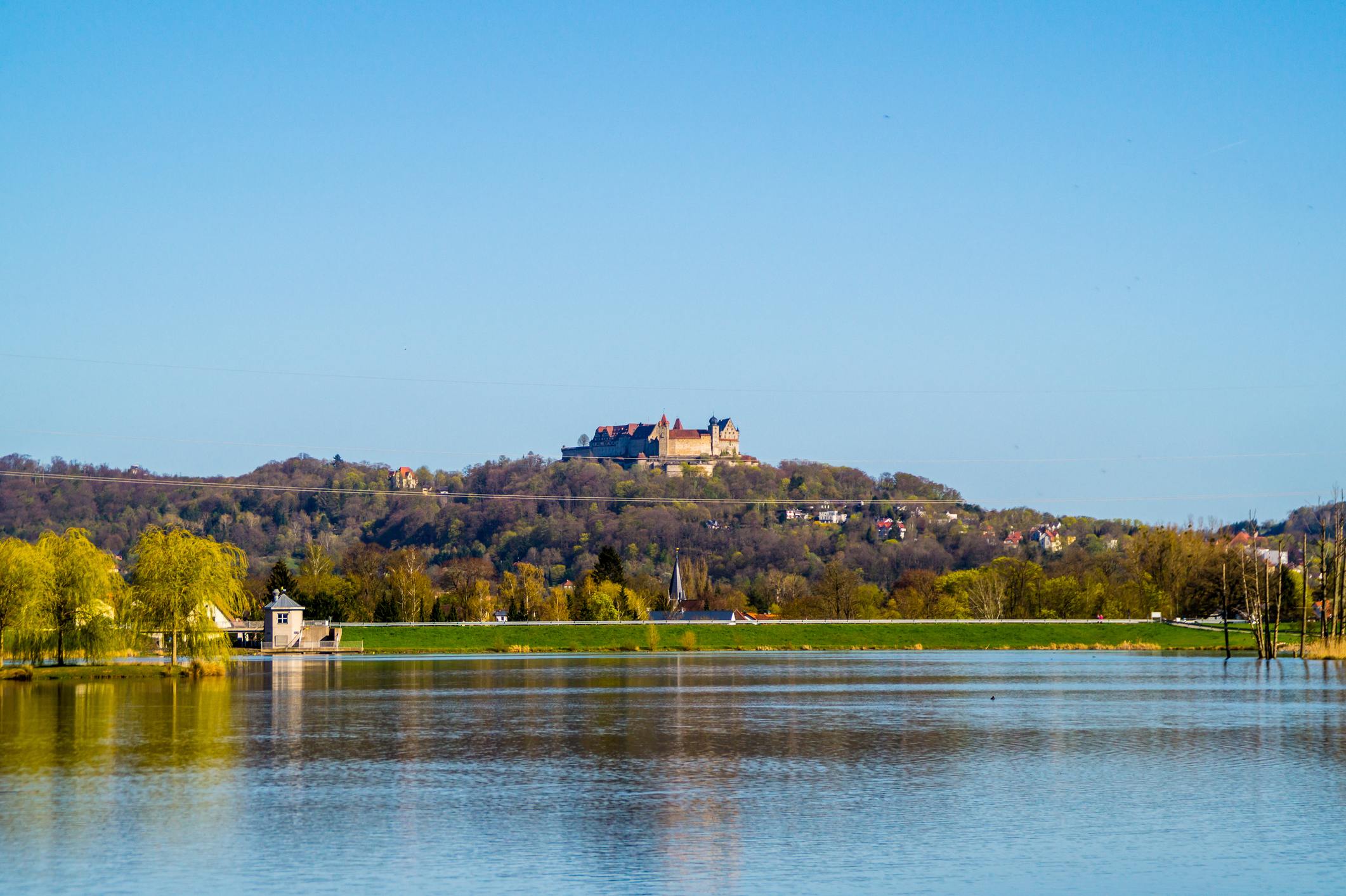 Ein großes historisches Schloss, bekannt als Coburg, thront auf einem baumbewachsenen Hügel unter einem klaren blauen Himmel, mit einem ruhigen See und kleinen Gebäuden im Vordergrund, umgeben von lebhaftem Frühlingsgrün. at Hochschule Coburg