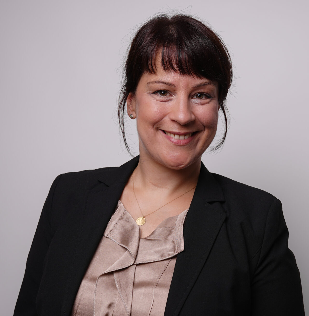 Stefanie Forkel Foto groß Stefanie Forkel, mit dunklem Haar in einem Pferdeschwanz, lächelt in die Kamera. Sie trägt einen schwarzen Blazer über einer beigen Seidenbluse und eine goldene Halskette. Der Hintergrund ist einfarbig und hell, was ihre Rolle an der Hochschule Coburg widerspiegelt. at Hochschule Coburg