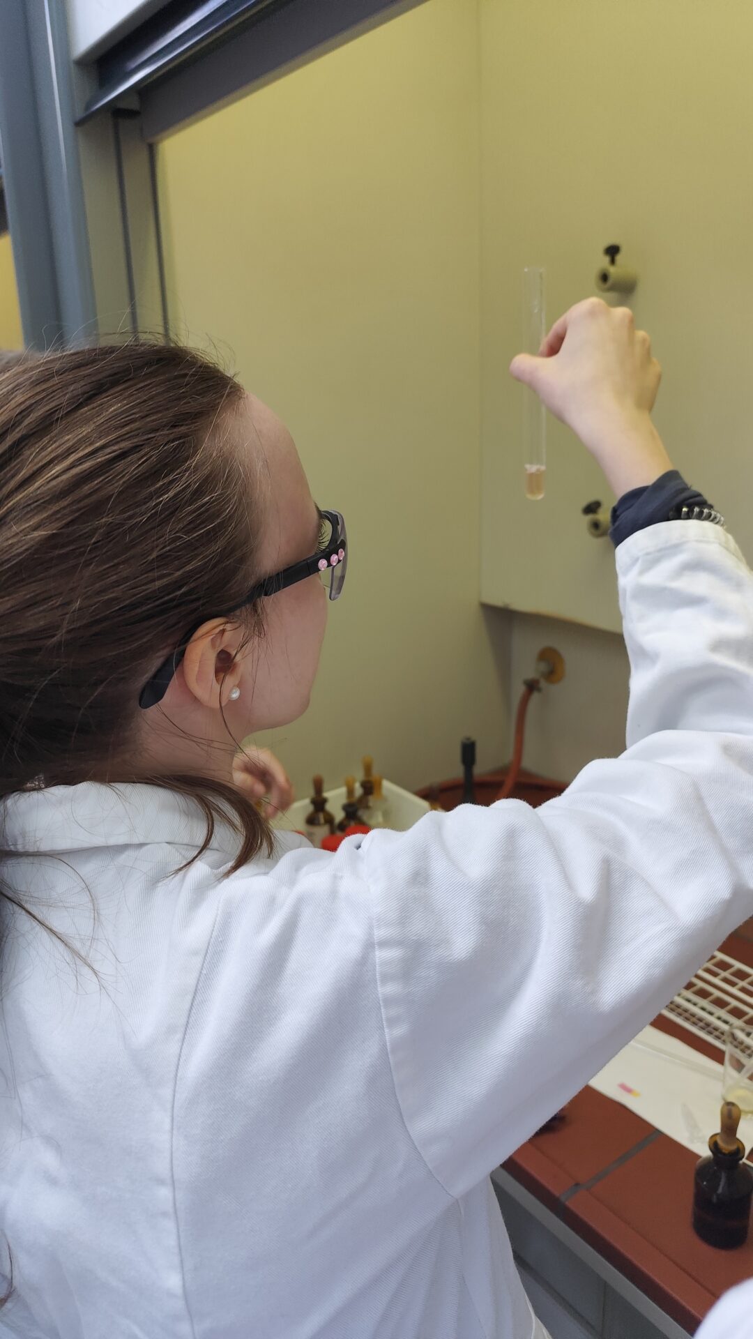 Eine Person in weißem Laborkittel und mit Brille hält ein Reagenzglas in einem Labor in der Nähe eines Abzugs hoch und untersucht dessen Inhalt während des Experimentiertags im Rahmen des Studieninfo-Programms auf dem Hochschul-Campus genau. at Hochschule Coburg