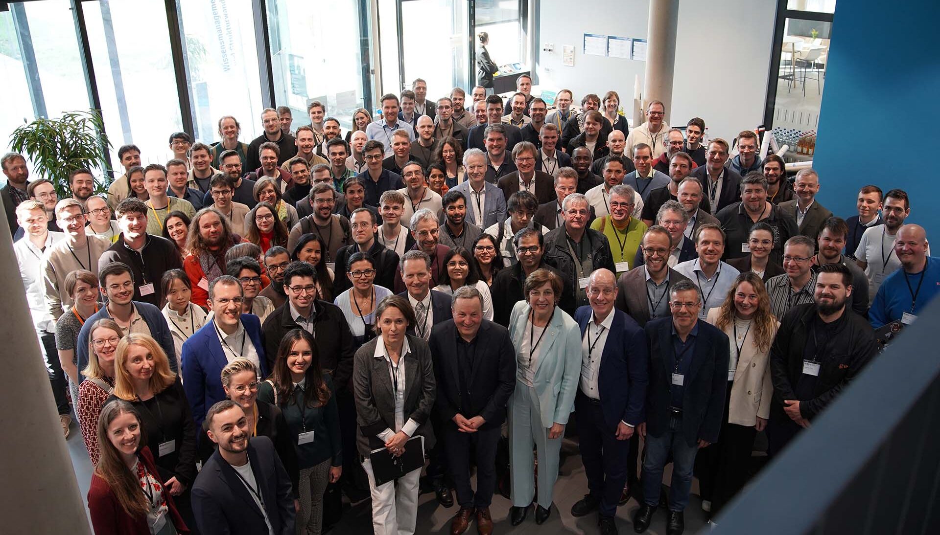 Eine große Gruppe von Doktorandinnen und Doktoranden, gekleidet in Geschäfts- oder Freizeitkleidung, steht in einem hellen, modernen Raum mit großen Fenstern in der Region eng beieinander, lächelt und blickt in die Kamera. at Hochschule Coburg