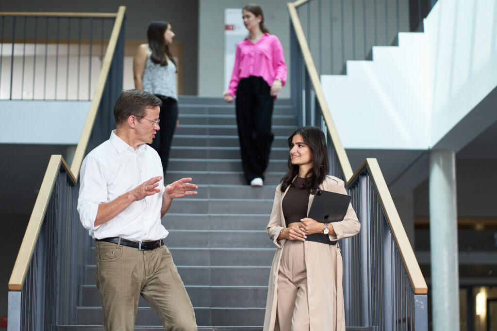 Two people have a social conversation while walking up stairs in a modern building, promoting health. The man in a white shirt gestures as he speaks, while the woman in beige holds a folder. Two other women walk down the stairs behind them. at Hochschule Coburg