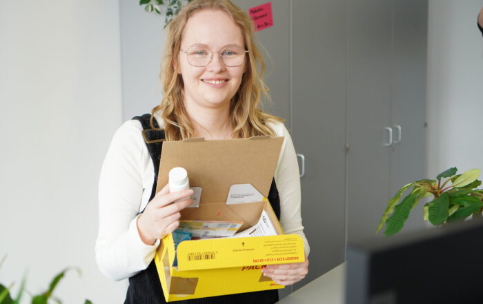 Eine lächelnde Person mit Brille hält ein geöffnetes gelbes Paket, das eine weiße Flasche und Papiere enthält. Sie steht in einem Büro, in dem im Hintergrund Schränke und eine Pflanze zu sehen sind. at Hochschule Coburg
