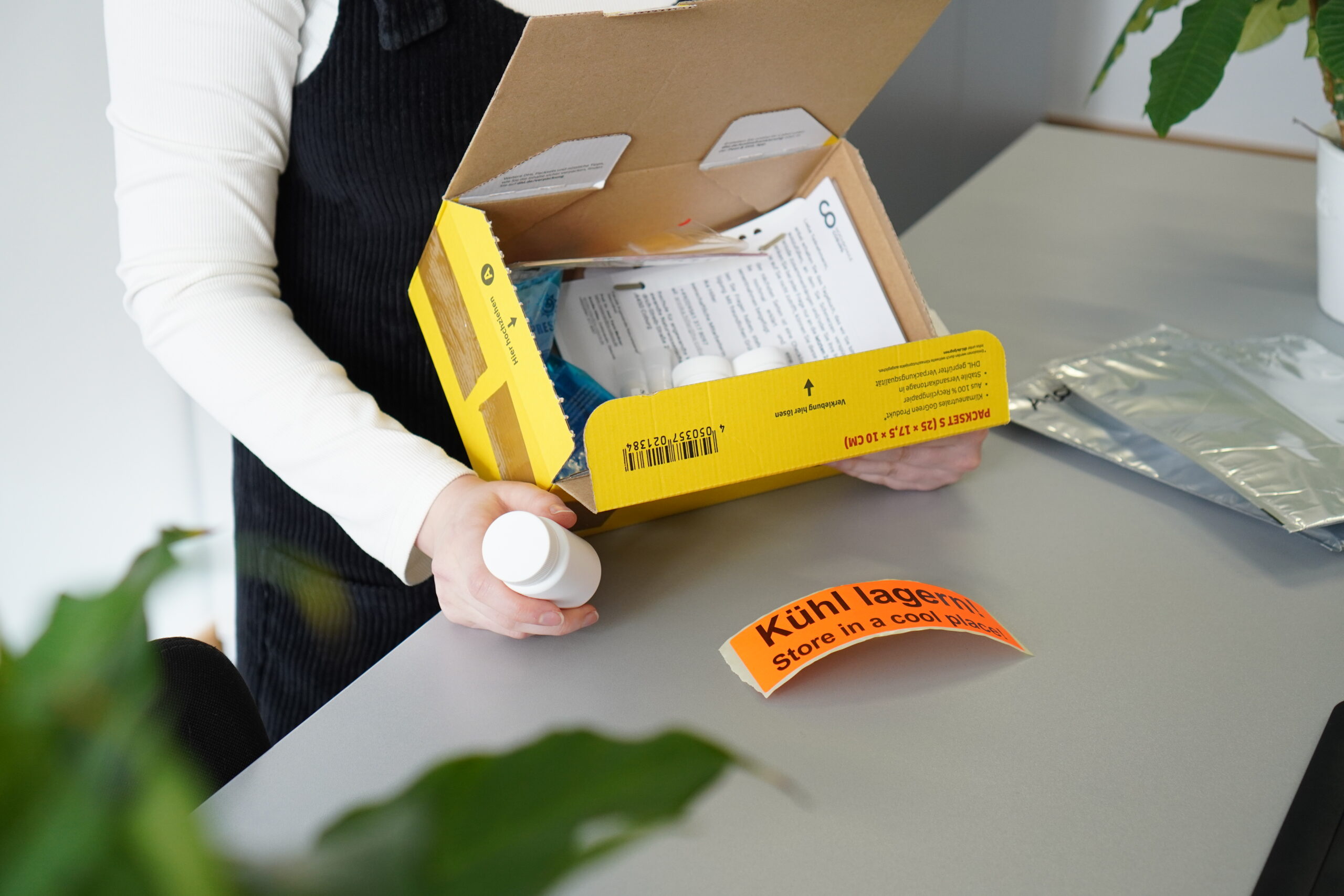 Eine Person öffnet eine gelbe Pappschachtel mit Medikamentenflaschen und Dokumenten - Elemente, die für die Gesundheit wichtig sind. Auf dem Tisch daneben liegt ein orangefarbenes Etikett mit der Aufschrift "Kühl lagern". at Hochschule Coburg