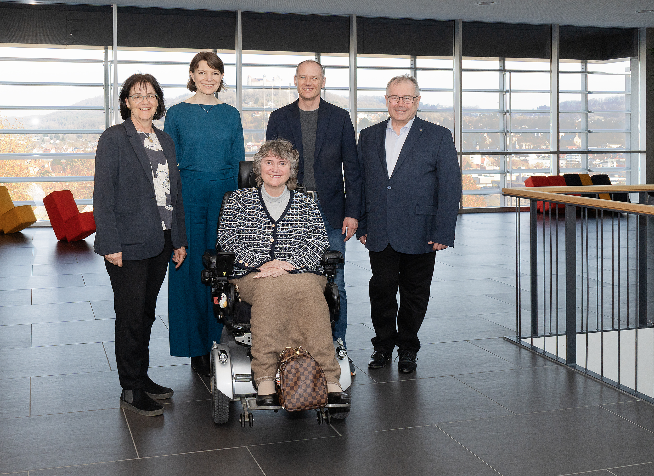 Fünf Erwachsene, darunter eine Frau im Rollstuhl, posieren und lächeln gemeinsam in einem hellen, modernen Raum mit großen Fenstern und Blick auf die Stadt und die Hügel - ein automatisch gespeicherter Entwurf von glücklichen Momenten. at Hochschule Coburg