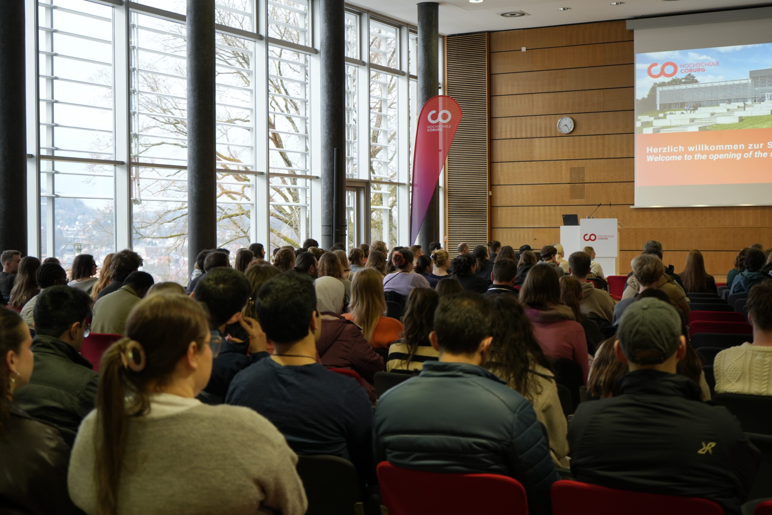 Eine große Gruppe von Menschen, darunter viele Erstsemester-Studis, sitzt in einem Konferenzraum und blickt auf einen Bildschirm mit einer Begrüßung in Deutsch und Englisch. Hohe Fenster lassen natürliches Licht herein, während ein Sprecher der Hochschule am Podium steht. at Hochschule Coburg
