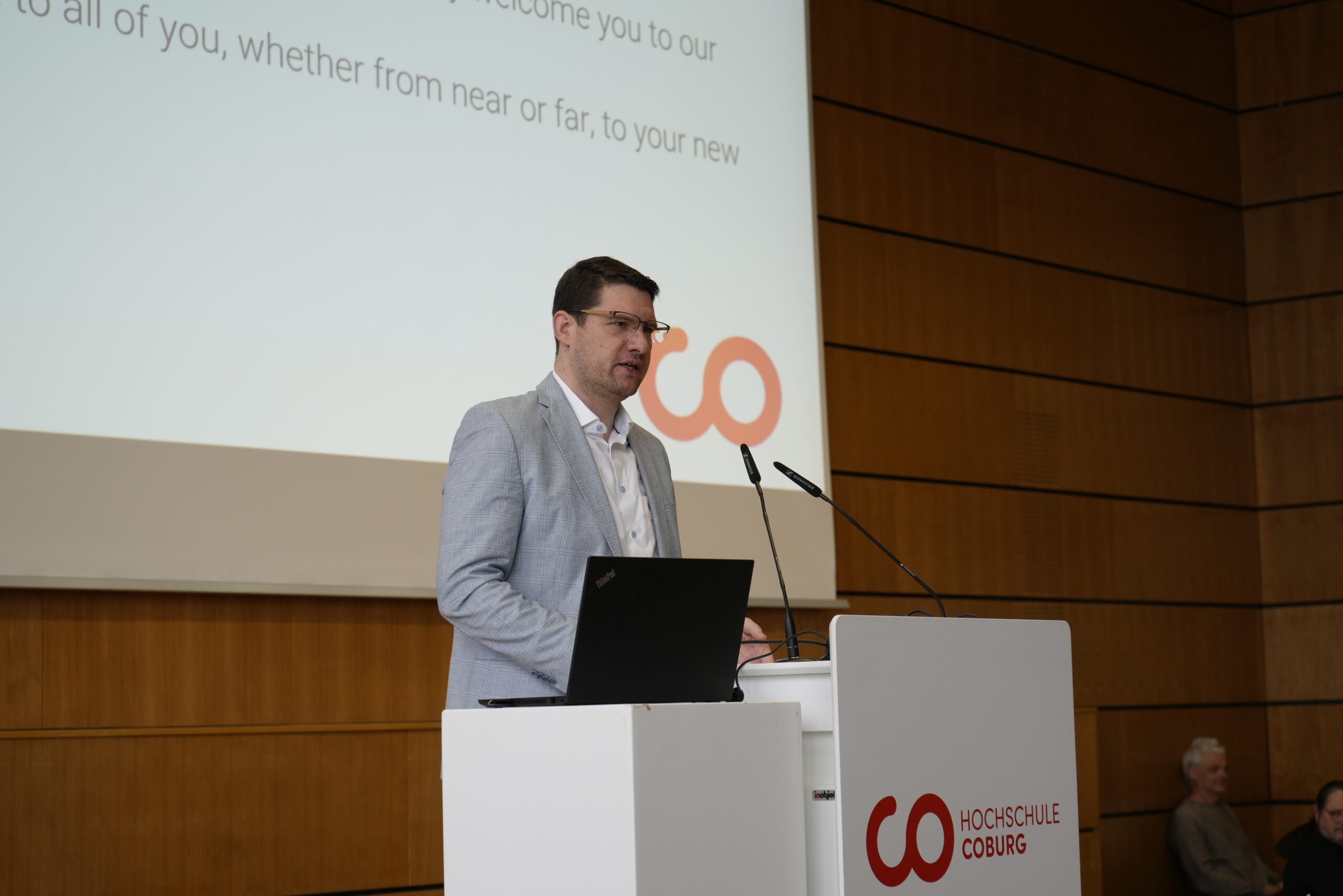 Ein Mann in einem hellgrauen Blazer spricht an einem Podium mit dem Logo der Hochschule Coburg zu den Erstsemesterstudenten in einem Hörsaal. Auf dem Podium steht ein Laptop, hinter ihm ist eine große Projektionsfläche mit Text zu sehen. at Hochschule Coburg