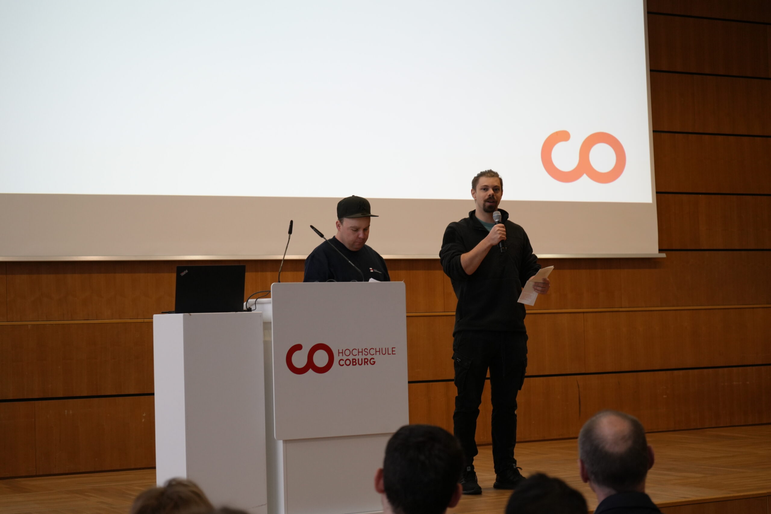 Zwei Personen stehen auf der Bühne an einem Podium, eine spricht in ein Mikrofon, während auf einer großen Leinwand das Logo der Hochschule Coburg erscheint. Im Vordergrund ist das Publikum zu sehen, darunter neugierige Erstsemester und Studis. at Hochschule Coburg