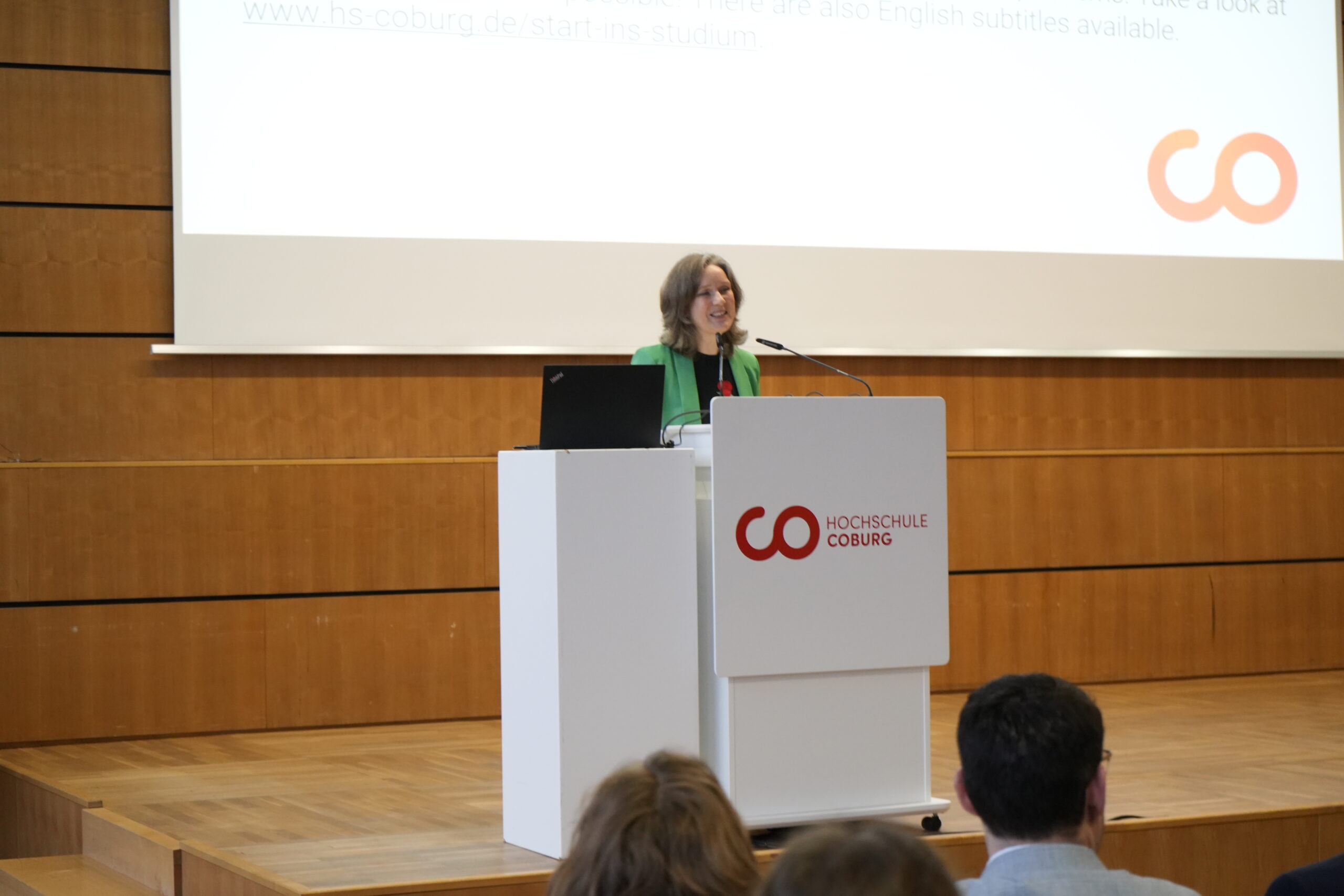 Eine Frau steht an einem Podium und hält einen Vortrag in einem Hörsaal, um das Erstsemester an der Hochschule Coburg zu begrüßen. Auf dem Podium und der Leinwand ist das Logo der Hochschule zu sehen, während mehrere Studis im Publikum aufmerksam zuhören. at Hochschule Coburg