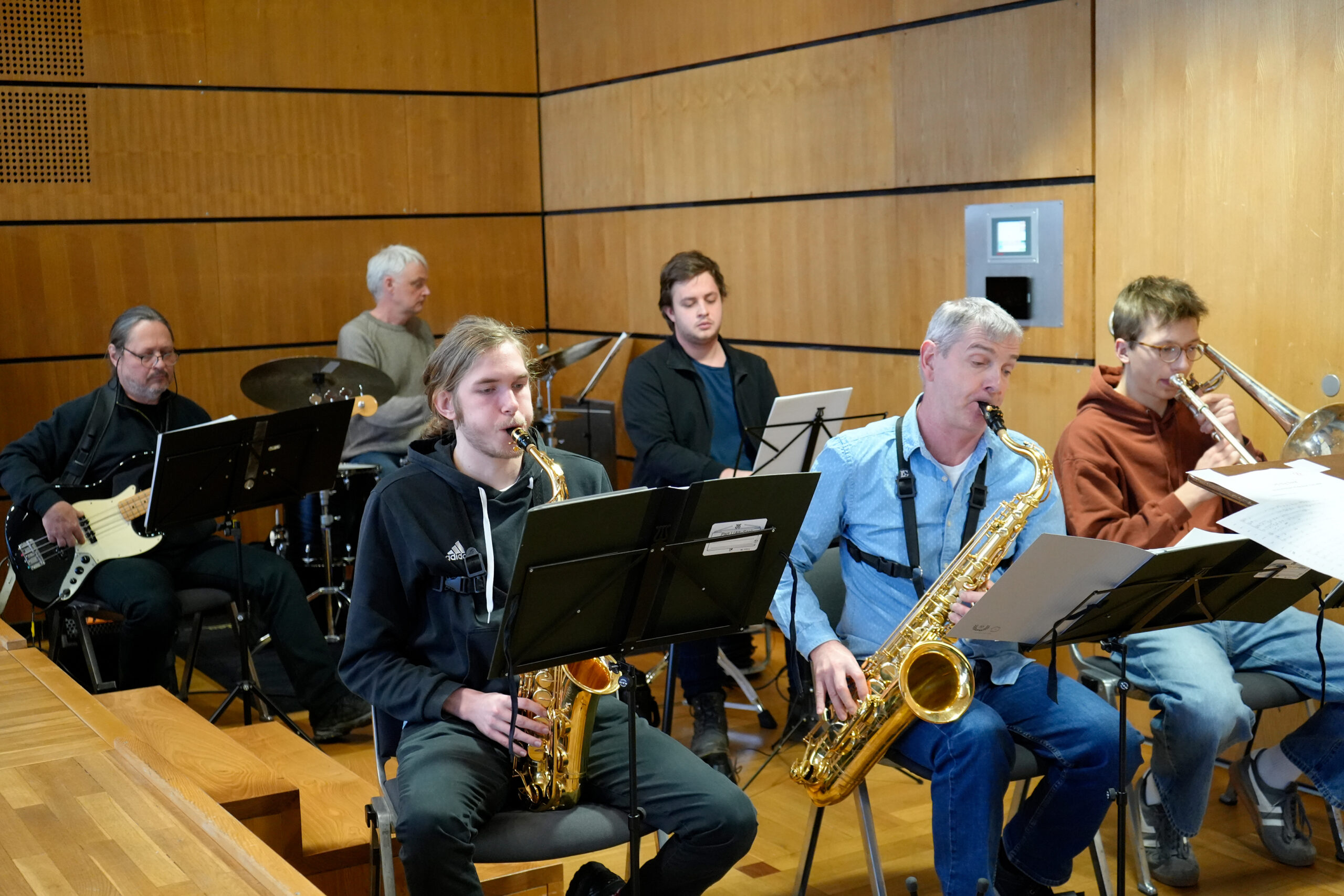 Sechs Musiker in einem holzgetäfelten Raum und spielen Saxophon, Posaune, Bassgitarre und Schlagzeug. Sie sitzen und konzentrieren sich auf Notenblätter und scheinen Teil einer Jazz- oder Konzertband zu sein. at Hochschule Coburg