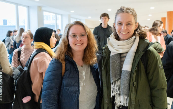 Zwei Frauen, beide mit Brillen und Winterjacken, lächeln sich in einem Innenraum der Hochschule an. Umgeben von anderen Erstsemestern und Studis in einem hell erleuchteten Flur, genießen sie eine lebhafte Gesprächsatmosphäre. at Hochschule Coburg