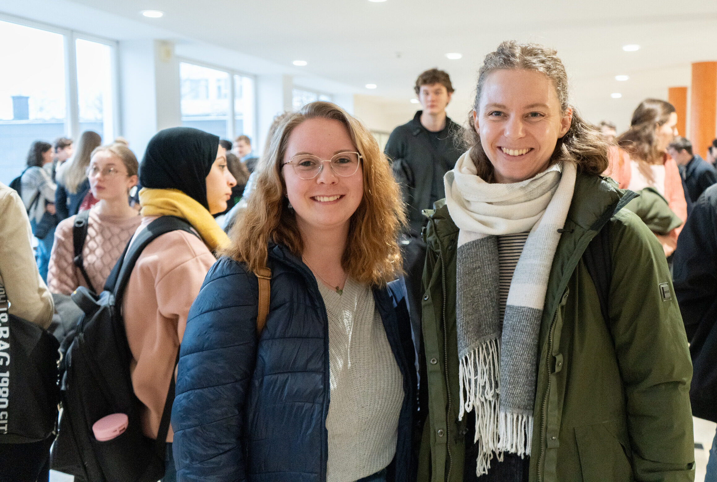 Zwei Frauen, beide mit Brillen und Winterjacken, lächeln sich in einem Innenraum der Hochschule an. Umgeben von anderen Erstsemestern und Studis in einem hell erleuchteten Flur, genießen sie eine lebhafte Gesprächsatmosphäre. at Hochschule Coburg