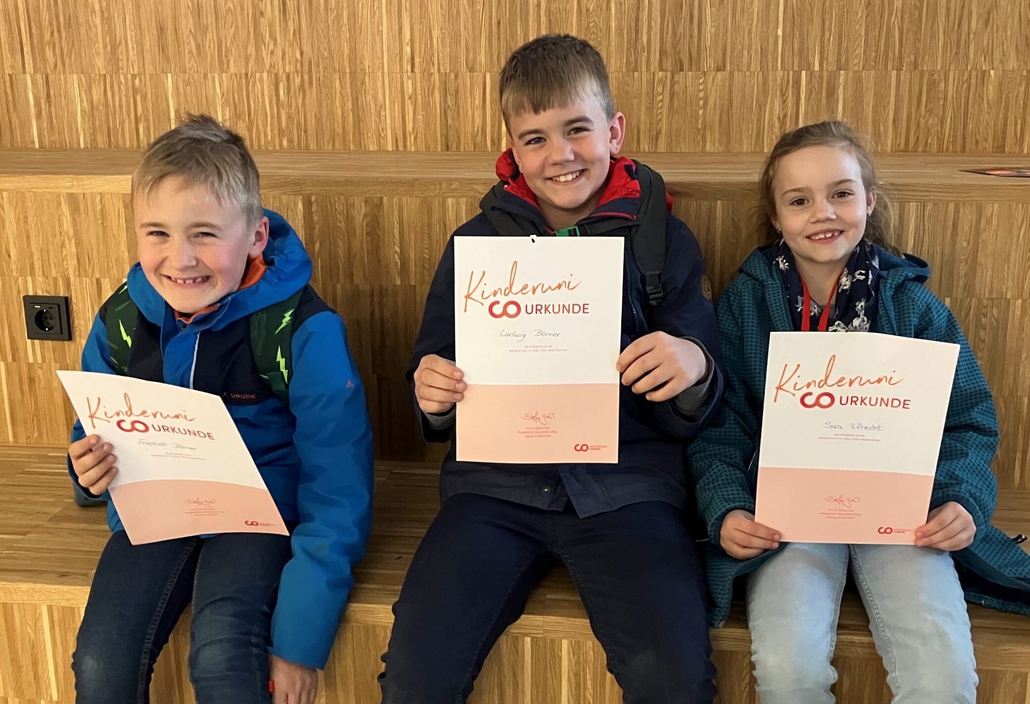 Drei lächelnde Kinder sitzen auf hölzernen Stufen und halten KinderUni-Urkunden mit der Aufschrift "CO Urkunde" in der Hand. Sie tragen Freizeitkleidung und Rucksäcke und sehen stolz und glücklich aus, nachdem sie gemeinsam Themen wie KI und Falschgeld erkundet haben. at Hochschule Coburg