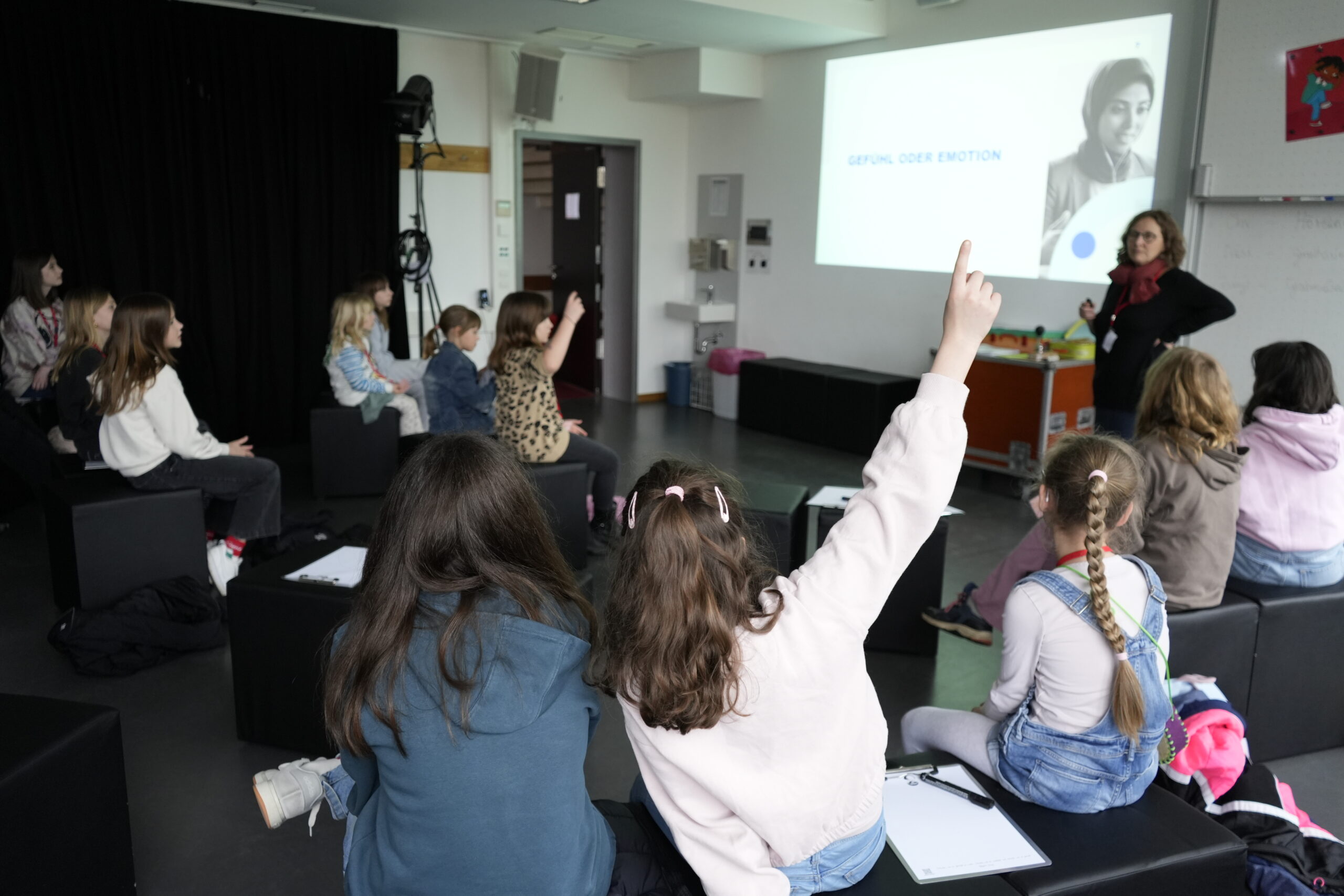 Eine Gruppe von Kindern sitzt auf schwarzen Sitzwürfeln in einem Klassenzimmer der KinderUni, mit Blick auf eine Lehrerin an der Stirnseite. Mehrere Kinder heben die Hände, als eine Präsentation über KI und Falschgeld mit Schwarz-Weiß-Fotos an die Wand projiziert wird. at Hochschule Coburg