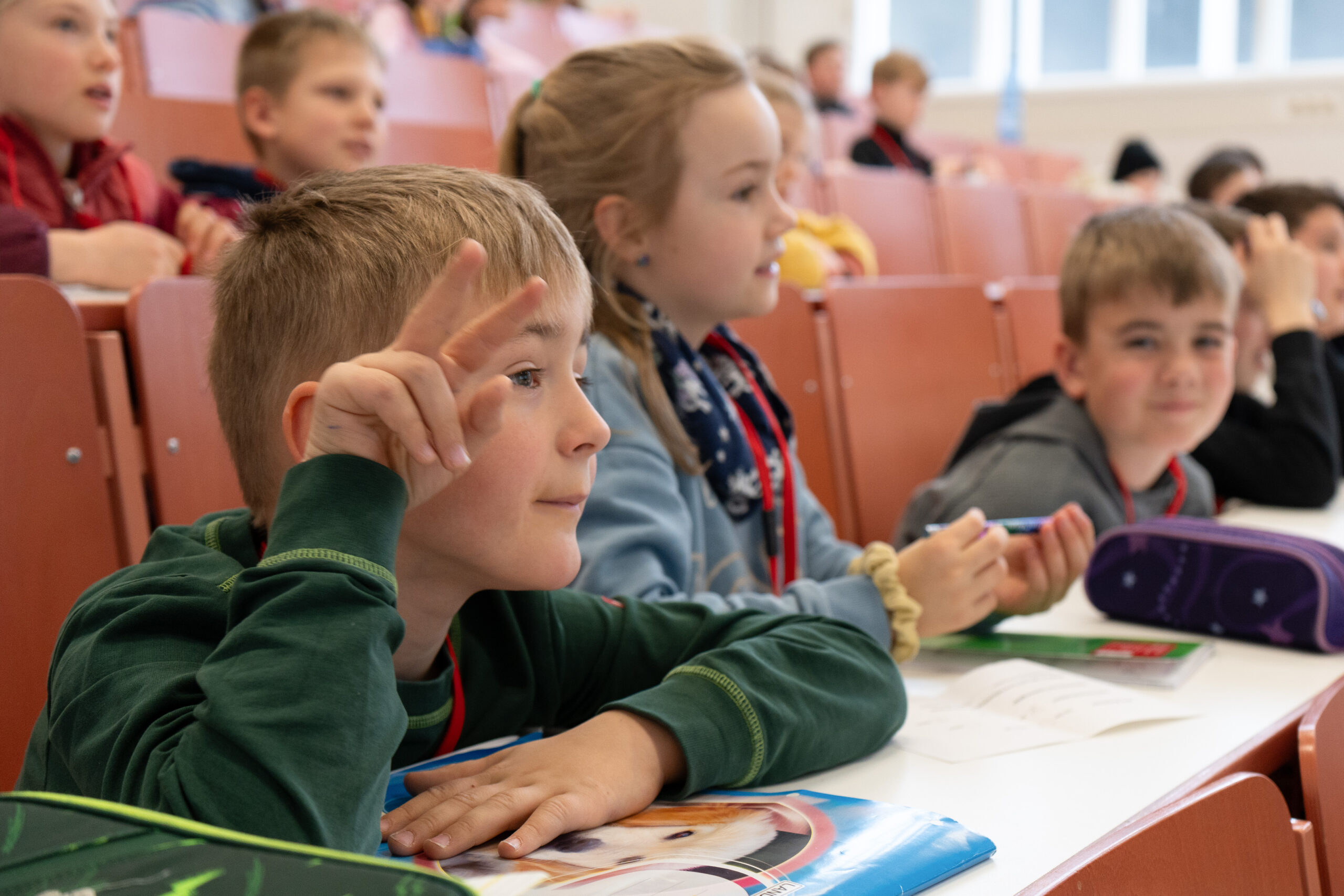 Eine Gruppe kleiner Kinder sitzt an den Tischen in einem Klassenzimmer der KinderUni. Ein Junge im Vordergrund hält zwei Finger hoch, als ob er eine Frage beantworten oder stellen würde. Die Kinder wirken aufmerksam und engagiert. at Hochschule Coburg