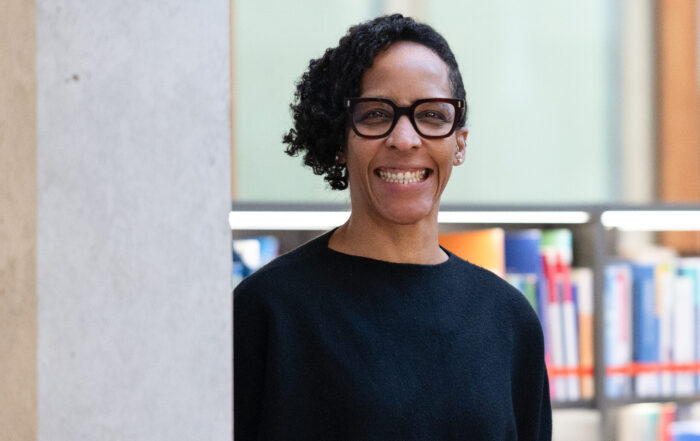 Eine Person mit kurzen lockigen Haaren und schwarzer Brille steht lächelnd in einem Innenraum an einer Wand, mit Bücherregalen und bunten Büchern im Hintergrund - vielleicht eine Professorin der Geburtshilfe, die ihr Wissen weitergibt. at Hochschule Coburg