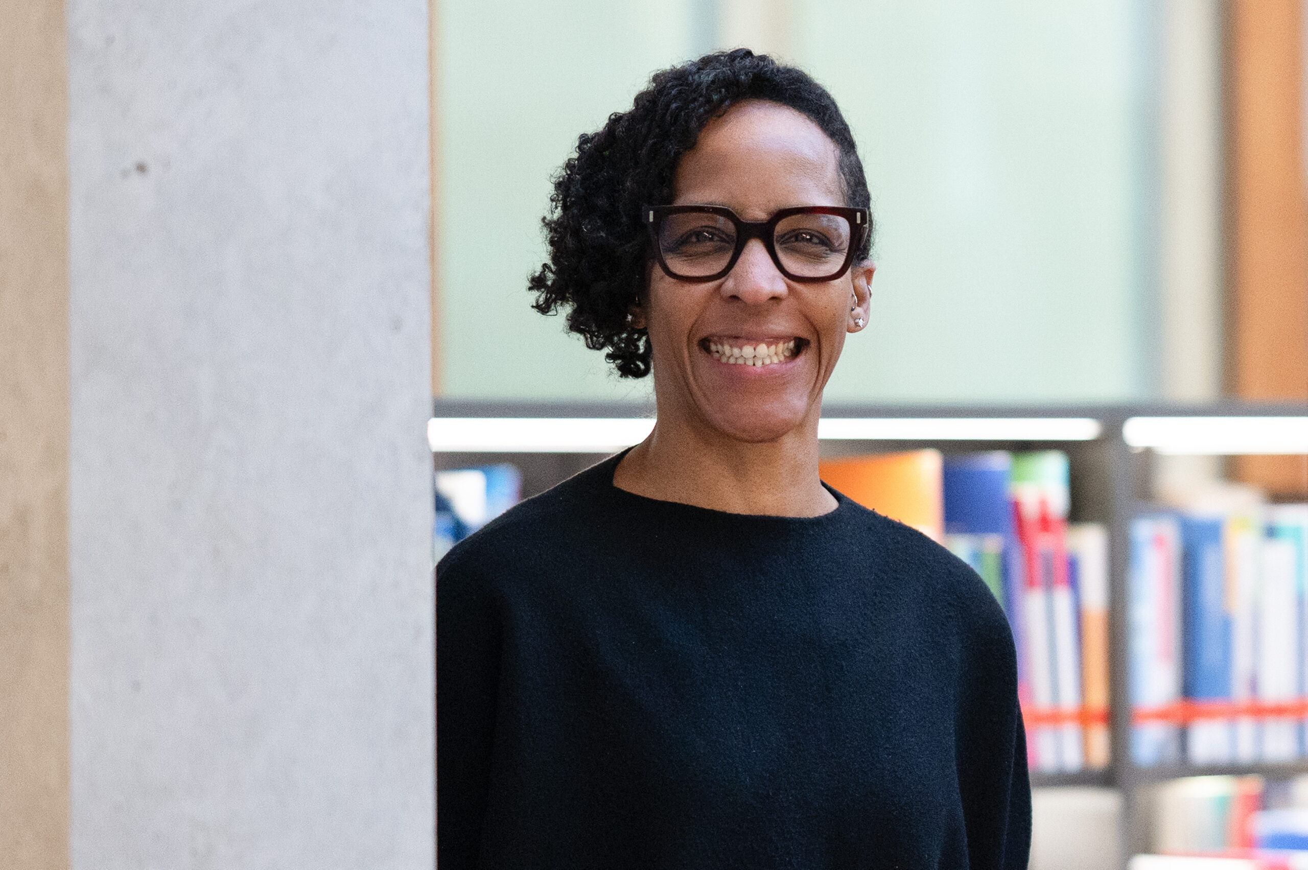 Eine Person mit kurzen lockigen Haaren und schwarzer Brille steht lächelnd in einem Innenraum an einer Wand, mit Bücherregalen und bunten Büchern im Hintergrund - vielleicht eine Professorin der Geburtshilfe, die ihr Wissen weitergibt. at Hochschule Coburg