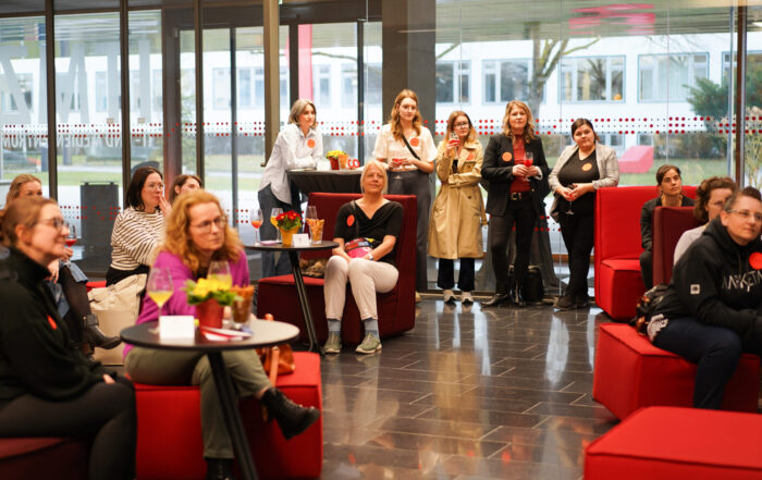 Eine Gruppe von Frauen sitzt und steht in einer modernen Lounge mit roten Stühlen während einer Gründerinnen-Auftaktveranstaltung und hört aufmerksam etwas aus dem Off zu. Einige halten Notizblöcke oder Getränke in der Hand, und mehrere tragen rote Kreisaufkleber. Im Hintergrund sind große Fenster zu sehen. at Hochschule Coburg