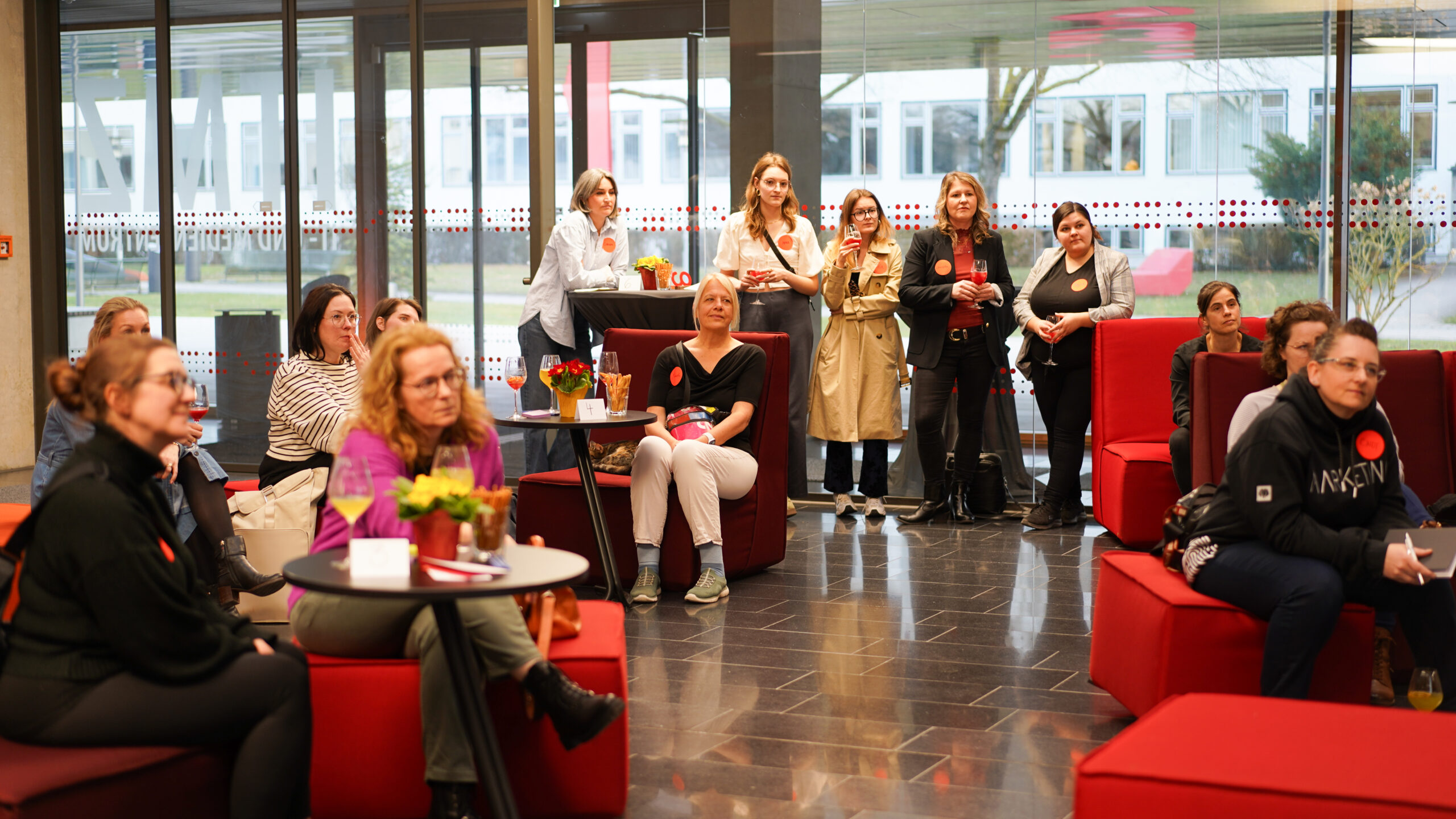 Eine Gruppe von Frauen sitzt und steht in einer modernen Lounge mit roten Stühlen während einer Gründerinnen-Auftaktveranstaltung und hört aufmerksam etwas aus dem Off zu. Einige halten Notizblöcke oder Getränke in der Hand, und mehrere tragen rote Kreisaufkleber. Im Hintergrund sind große Fenster zu sehen. at Hochschule Coburg