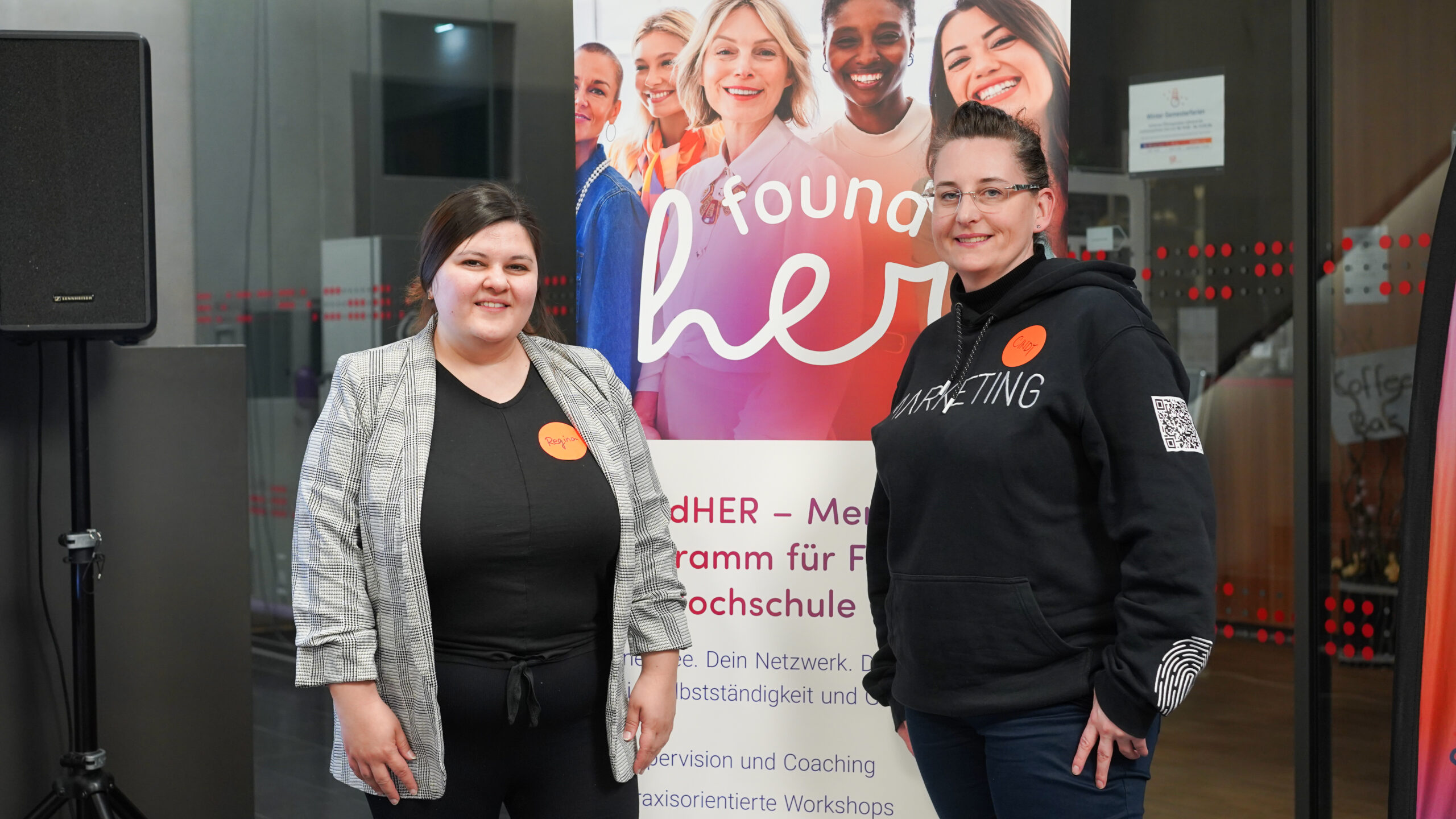 Zwei Frauen lächeln vor einem "foundHER"-Banner bei der Kick-off-Veranstaltung des Programms. Das Banner mit verschiedenen Frauengesichtern und deutschem Text wirbt für die zweite Runde von foundHER für Frauen an einer Universität. Beide tragen Freizeit- und Business-Kleidung. at Hochschule Coburg