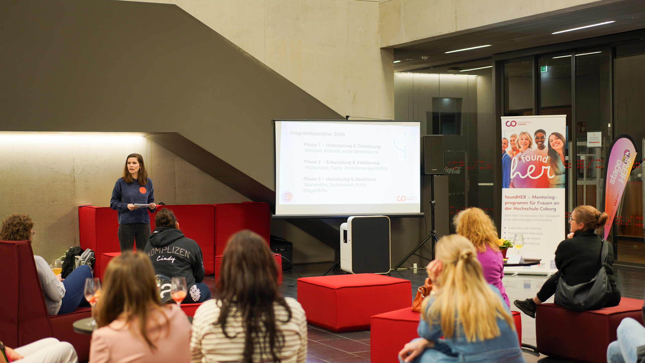 Eine Frau steht neben einem Projektor und hält vor einem sitzenden Publikum in einem modernen Innenraum eine Gründungspräsentation. Im Hintergrund sind ein Banner und Informationsmaterial zu sehen. at Hochschule Coburg