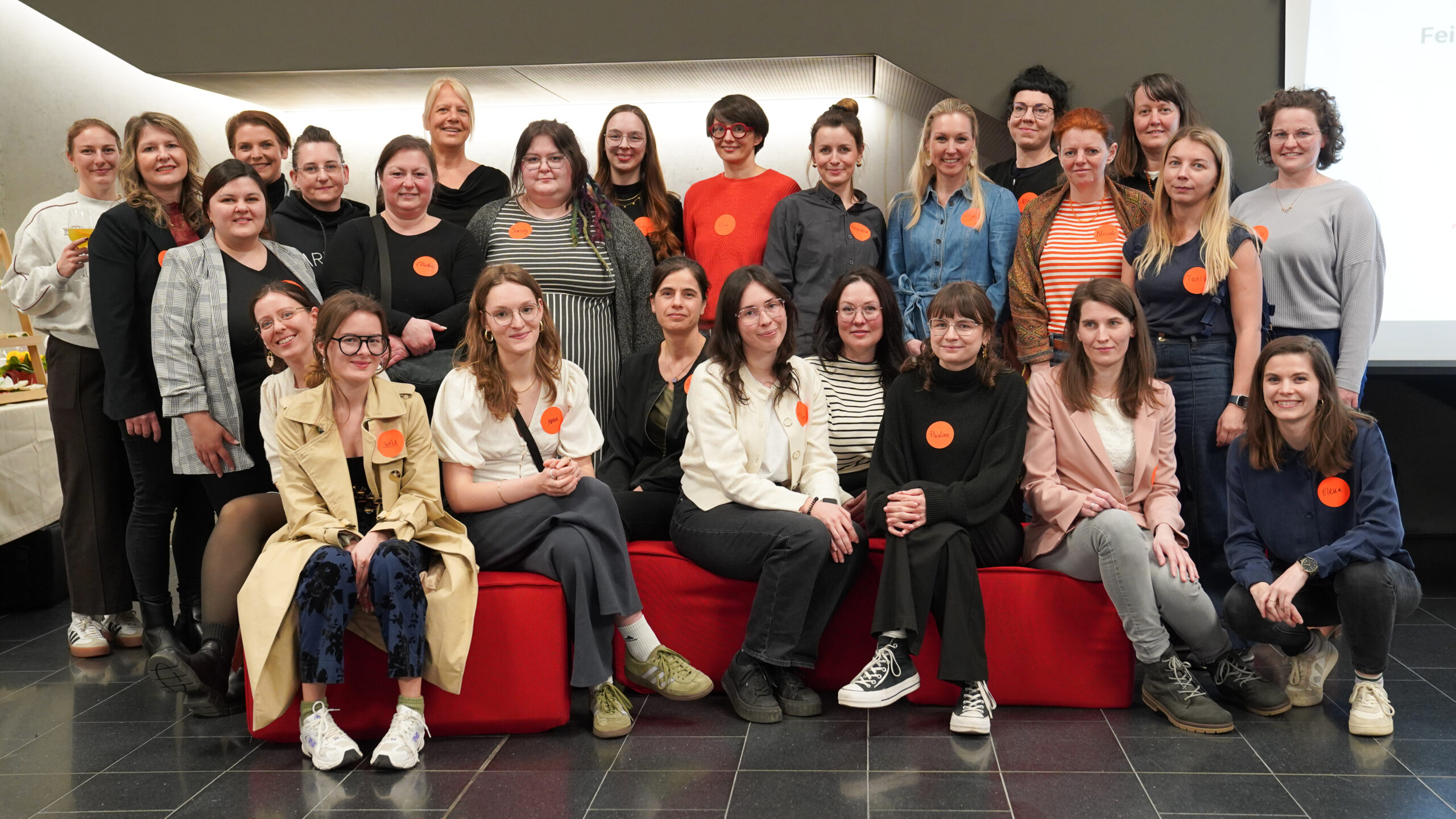 Eine Gruppe von 26 Frauen posiert gemeinsam in einem Innenraum beim Gründungsauftakt für die zweite Runde, viele tragen große orangefarbene Kreisaufkleber. Einige sitzen auf einer roten Bank, während andere hinter ihnen stehen, alle lächeln für das Foto. at Hochschule Coburg
