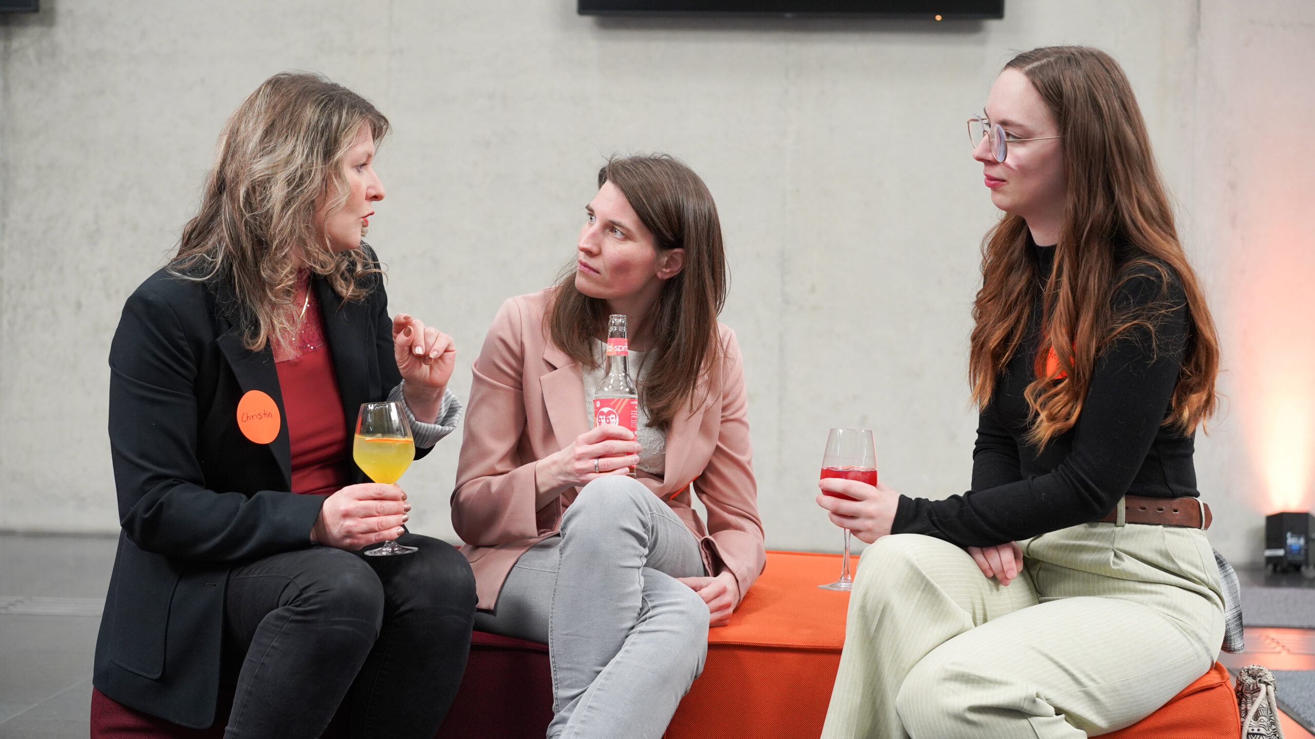 Drei Frauen sitzen auf orange- und bordeauxfarbenen Stühlen zusammen und unterhalten sich beim Kick-off von foundHER. Jede hält ein Getränk in der Hand, zwei tragen orangefarbene Namensschilder. Der Hintergrund ist eine helle, unifarbene Wand. at Hochschule Coburg