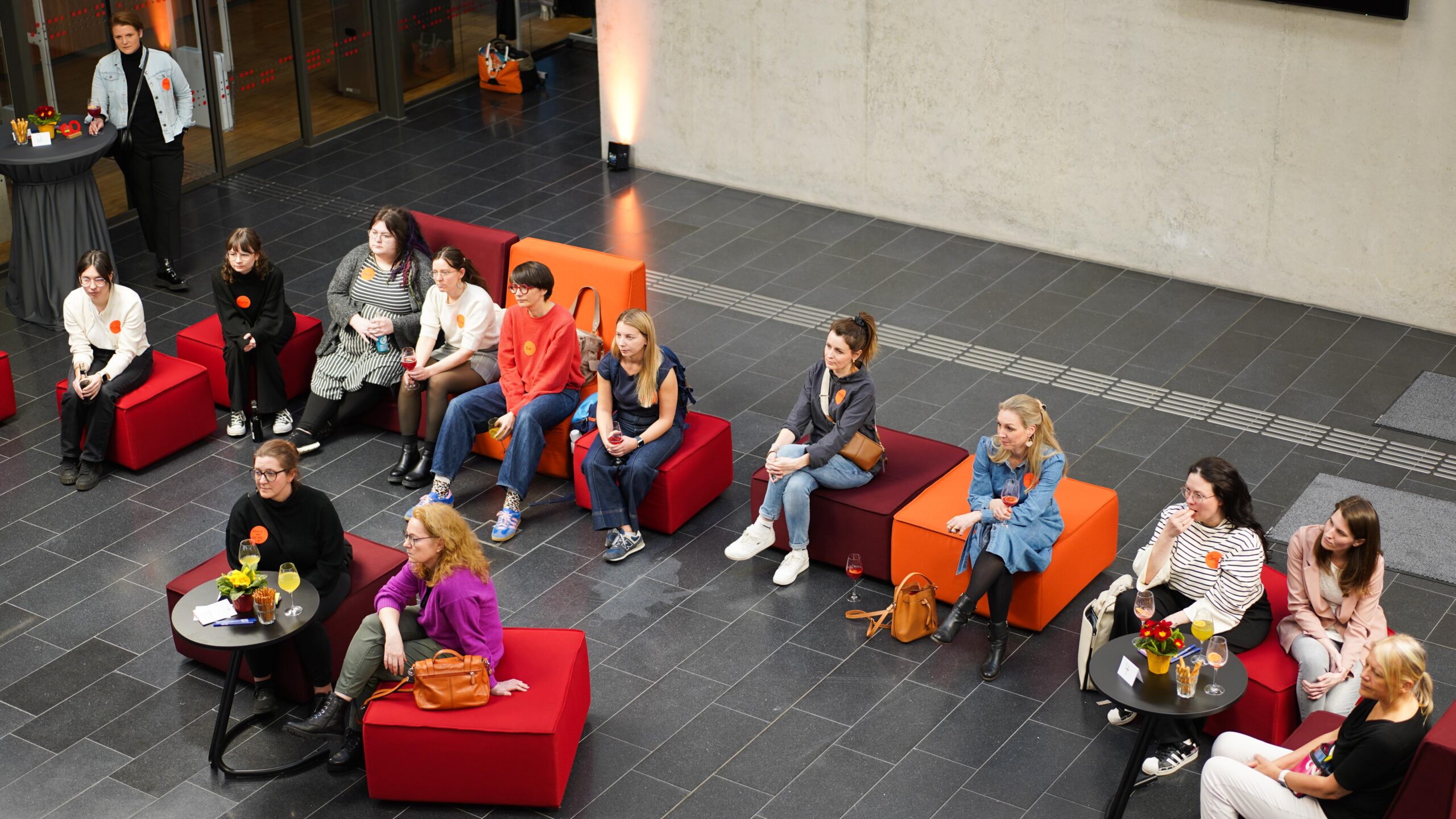 Eine Gruppe von Menschen sitzt auf roten Stühlen und schwarzen Tischen in einem modernen Innenraum während des FoundHER Kick-offs und beobachtet aufmerksam etwas im Off. Einige halten Getränke in der Hand, und auf den Tischen stehen Blumen für die zweite Runde. at Hochschule Coburg