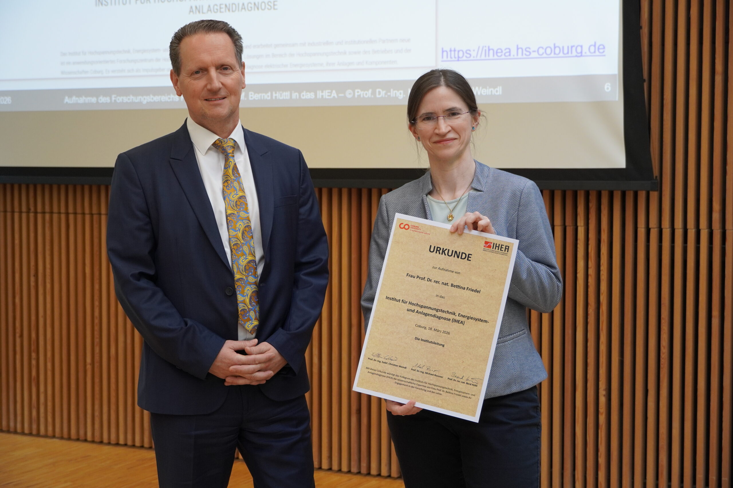 Ein Mann im Anzug und eine Frau mit einer Urkunde stehen nebeneinander und posieren für ein Foto in einem Innenraum mit einer Holzwand und einer projizierten Präsentationswand im Hintergrund. at Hochschule Coburg