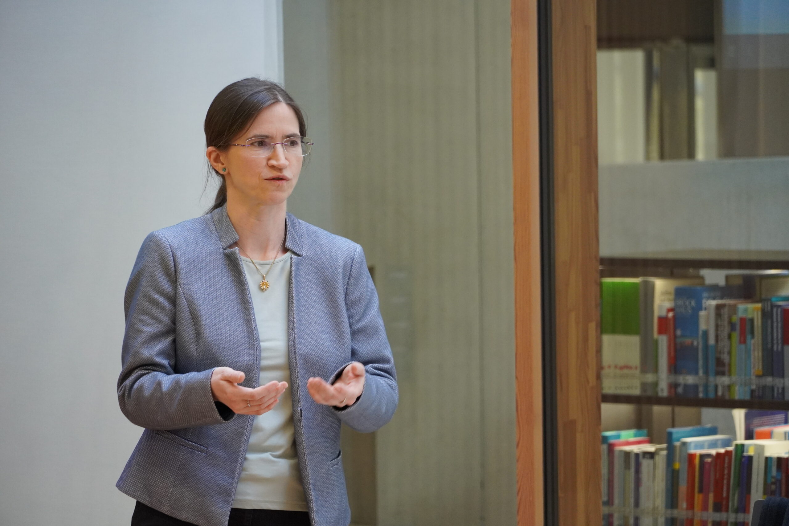 Eine Frau, die eine Brille und einen grauen Blazer trägt, gestikuliert mit den Händen, während sie spricht. Hinter ihr ist eine Glaswand mit Regalen voller Bücher zu sehen. at Hochschule Coburg